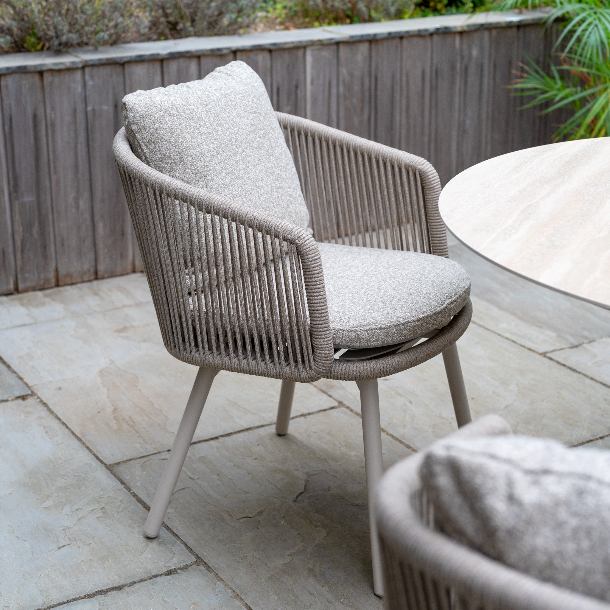 The Theia 4 Seat Round Rope Dining Set with Albatross Table in Oatmeal is arranged on a tiled patio, featuring beige cushioned chairs beside a round stone table.