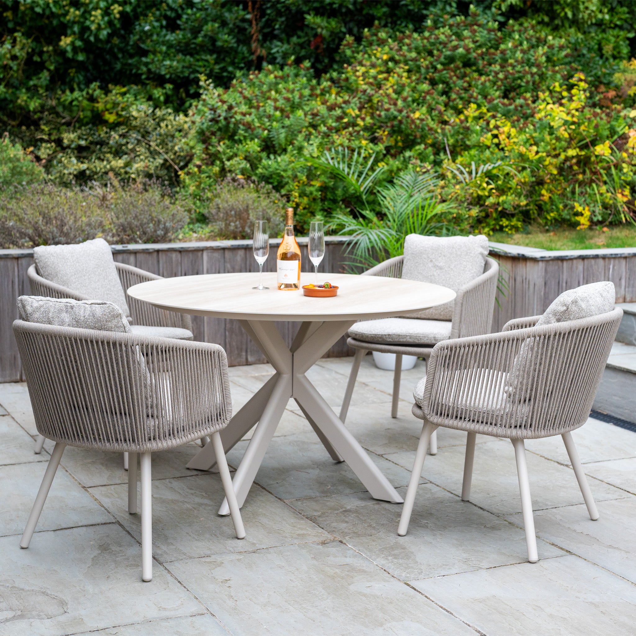 The Theia 4 Seat Round Rope Dining Set with Albatross Table in Oatmeal is arranged outdoors, surrounded by greenery—ideal for stylish alfresco dining with wine, glasses, and snacks.