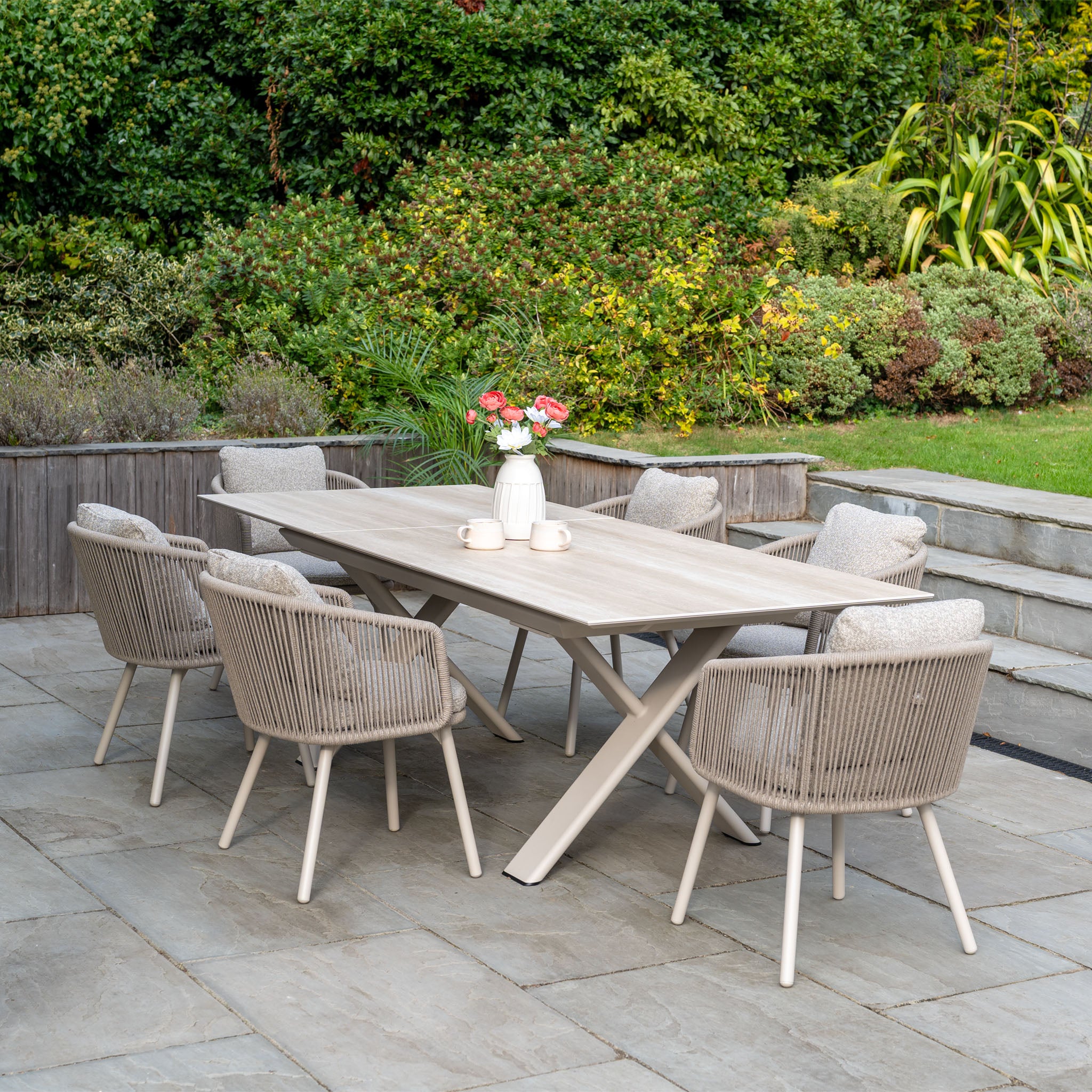 Outdoor patio featuring the Theia 6 Seat Rope Dining Set with Albatross Extending Table in Oatmeal and a vase of flowers, surrounded by greenery—ideal for outdoor dining.