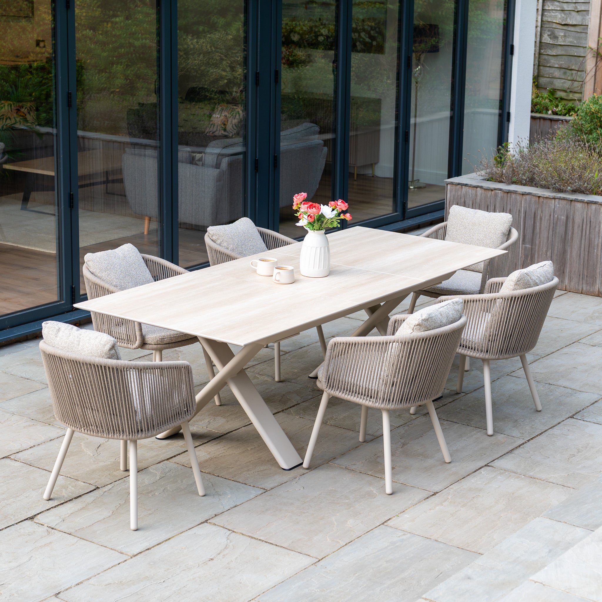 Theia 6 Seat Rope Dining Set with Albatross Extending Table in Oatmeal, plus six cushioned chairs, a flower vase, and mugs arranged on a stone patio.