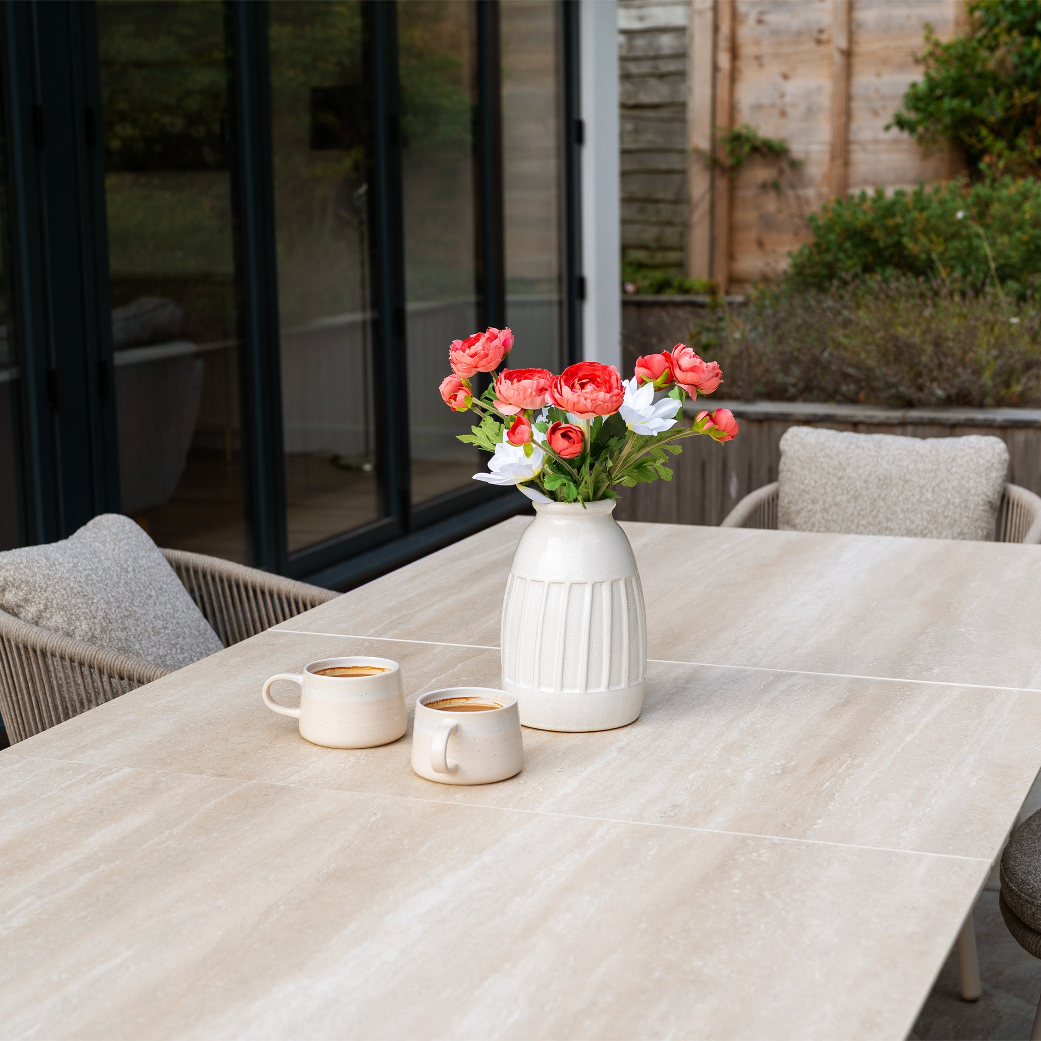 A ceramic vase with pink flowers and two coffee cups sit on the Albatross Extending Table, surrounded by cushioned chairs from Theia 6 Seat Rope Dining Set in Oatmeal.