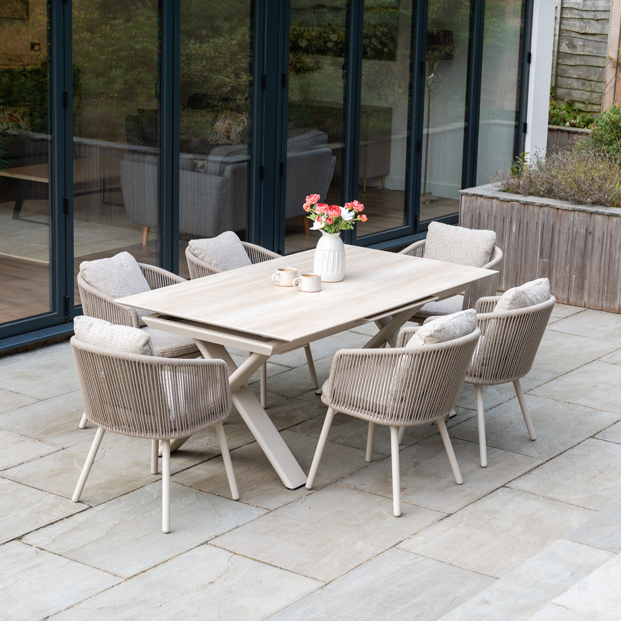 Theia 6 Seat Rope Dining Set with Albatross Extending Table in Oatmeal, arranged with a vase of flowers on a stone patio.