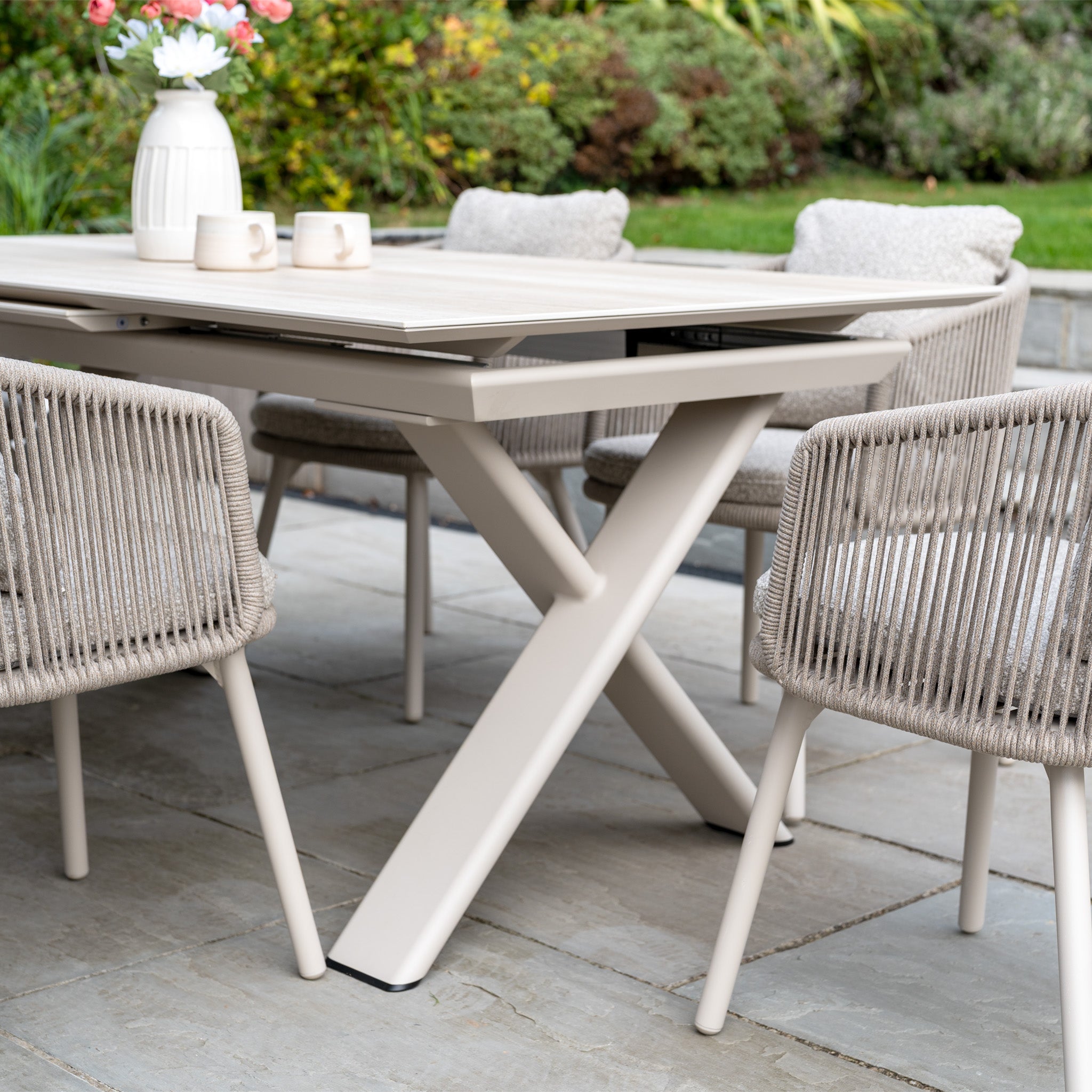 Theia 6 Seat Rope Dining Set with Albatross Extending Table in Oatmeal, featuring a vase of flowers on the patio with a garden view.