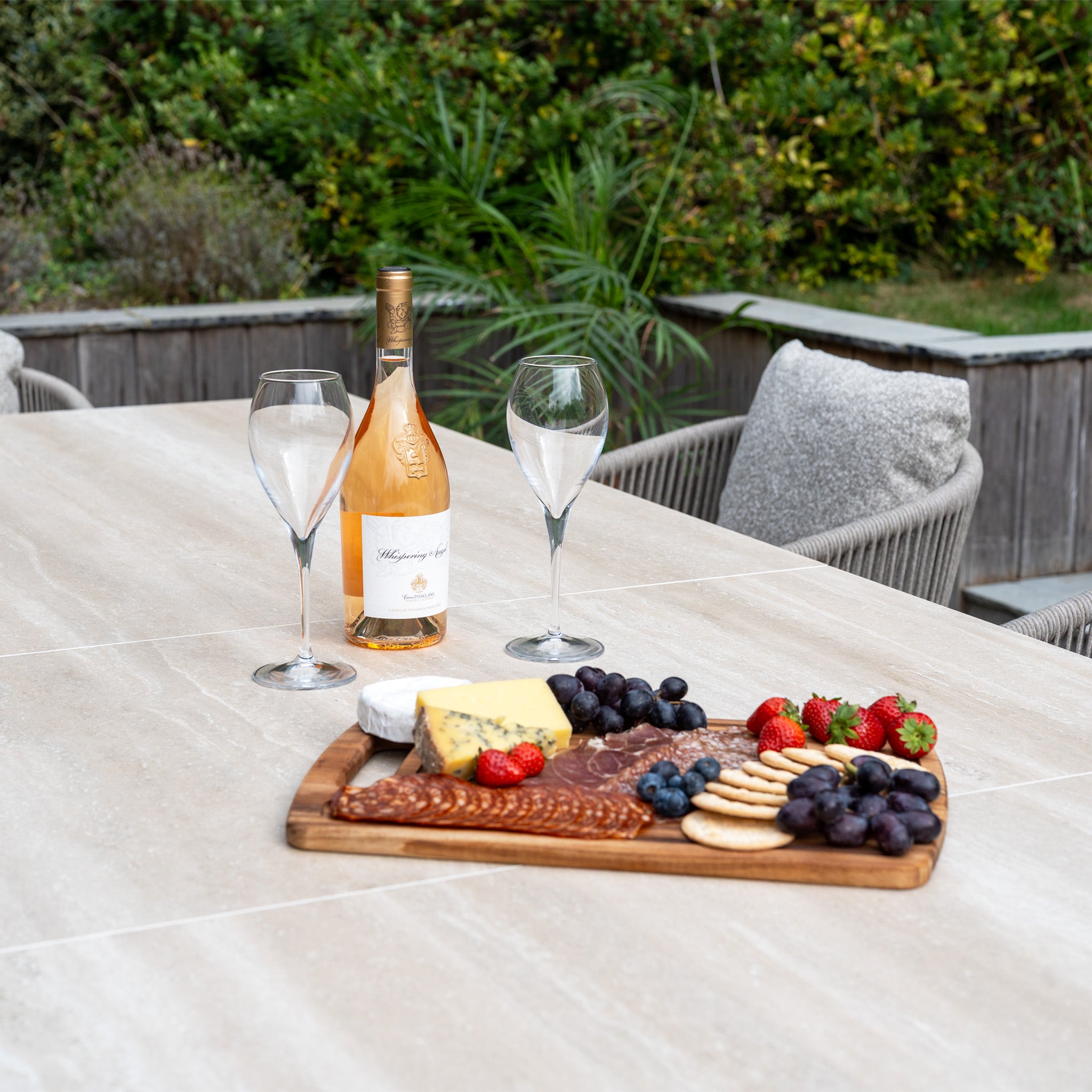 A bottle of wine, two glasses, and a cheese platter sit on the Theia 8 Seat Rope Dining Set with Albatross Extending Table in Oatmeal, arranged outdoors against a lush garden backdrop.