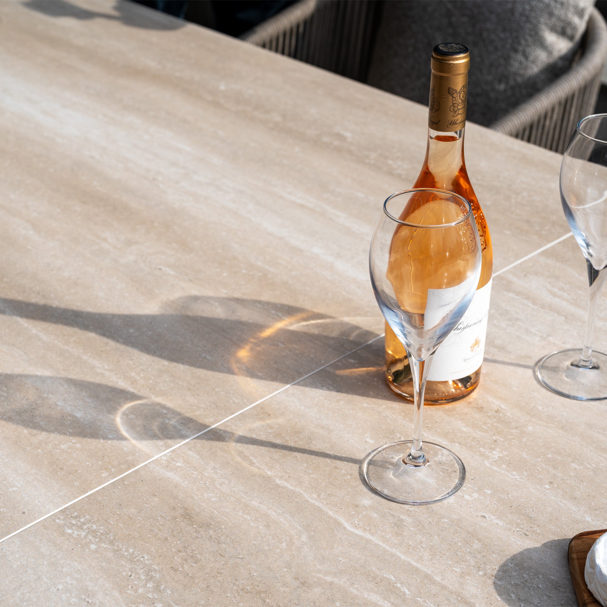 A bottle of rosé wine and two empty wine glasses rest on a beige stone table from the Theia 8 Seat Rope Dining Set with Albatross Extending Table in Oatmeal, bathed in warm sunlight.