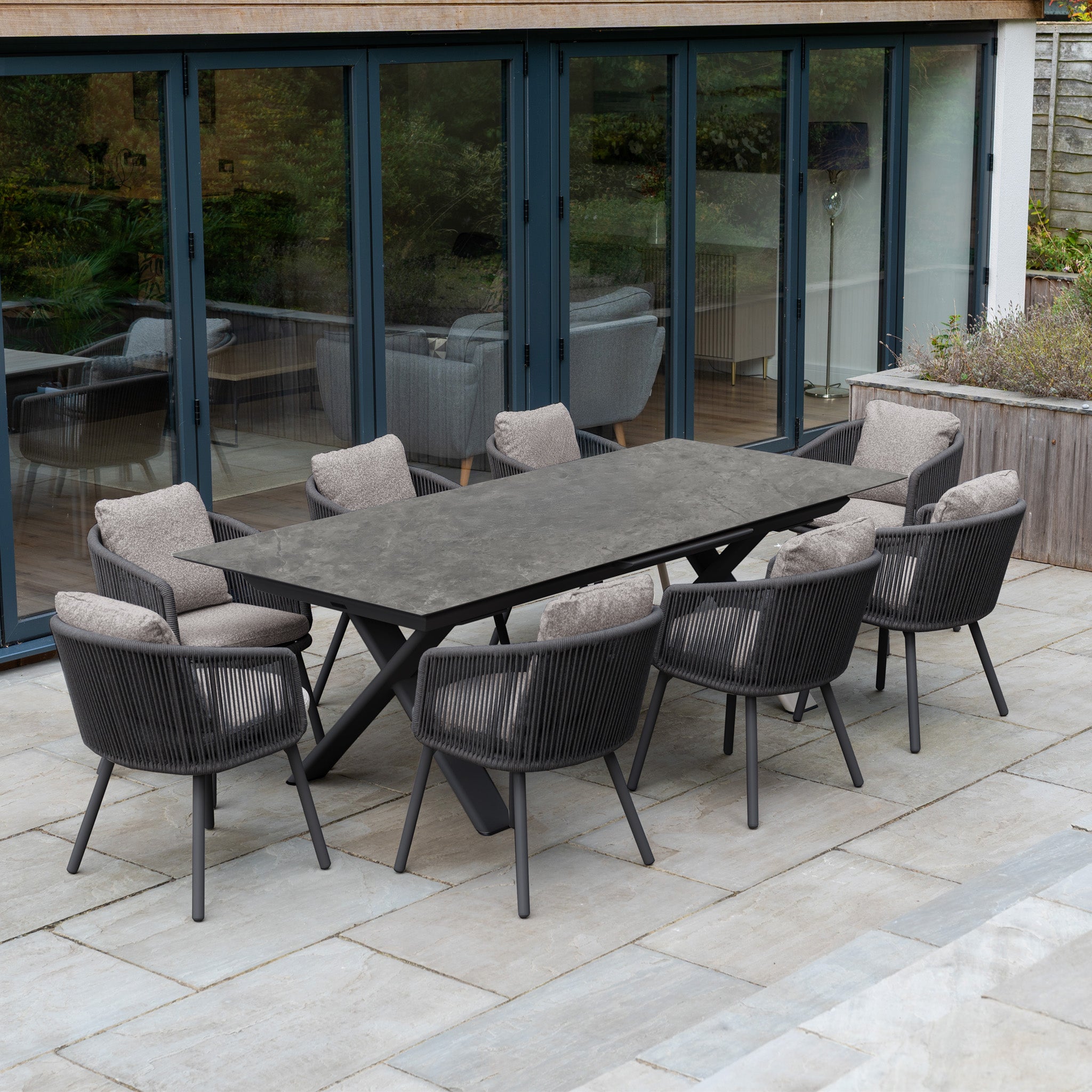 Theia 8 Seat Rope Dining Set with Albatross Extending Table in Truffle, featuring cushioned chairs, sits on a stone patio near glass doors.