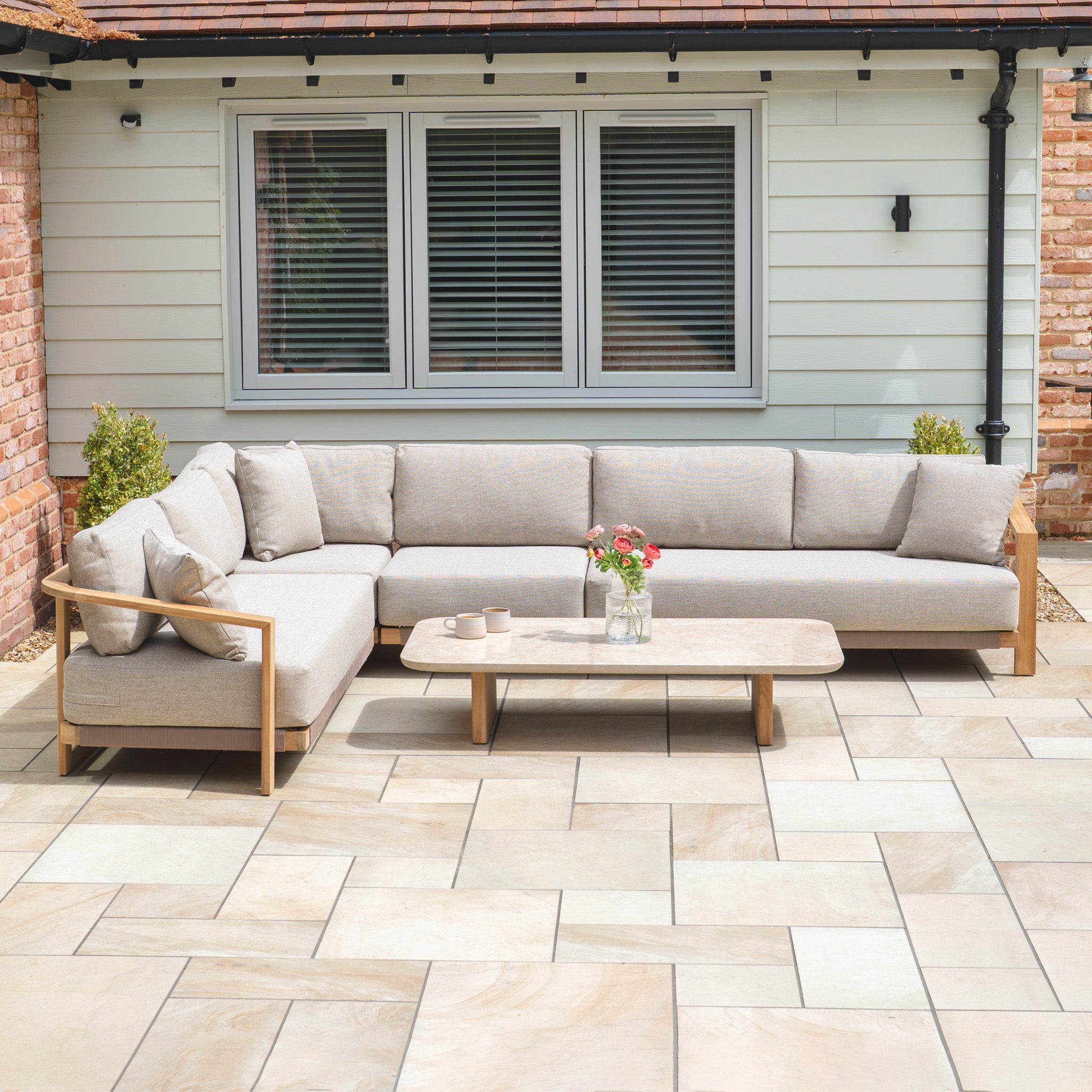 Outdoor patio with a Tivoli Large Corner Set with Marble Coffee Table in Fawn, featuring a beige sectional sofa and marble table accented by a flower vase in front of a house window.