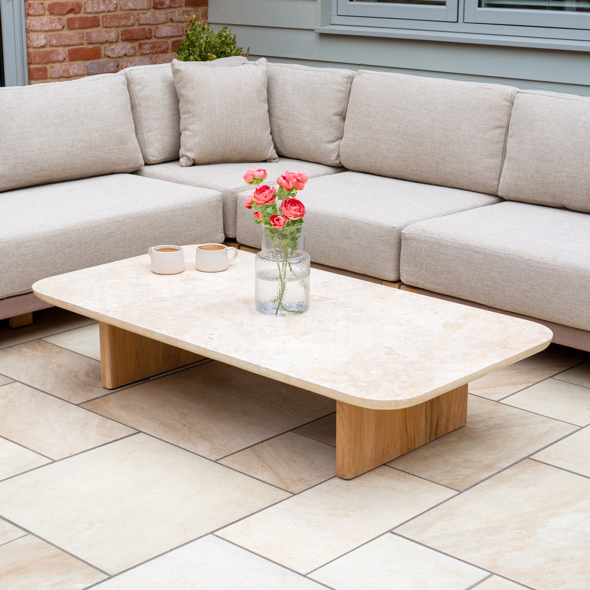 The Tivoli Large Corner Set with Marble Coffee Table in Fawn is displayed on a tiled patio, featuring pink flowers in a jar.