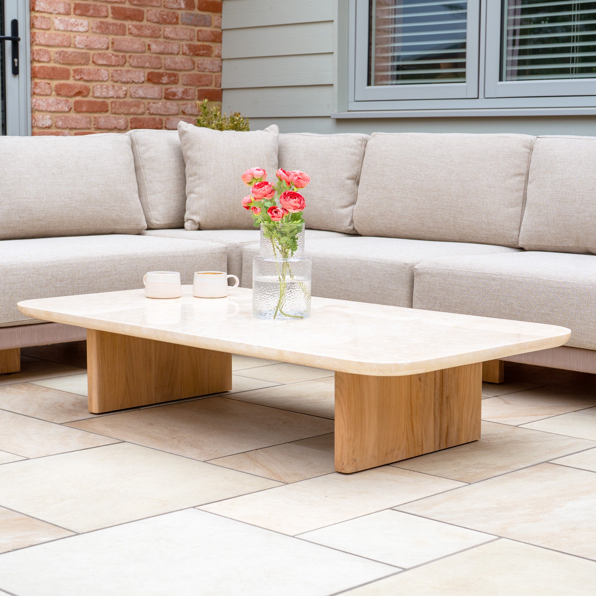 The Tivoli Large Corner Set with Marble Coffee Table in Fawn is displayed on a tiled patio, styled with flowers and mugs.