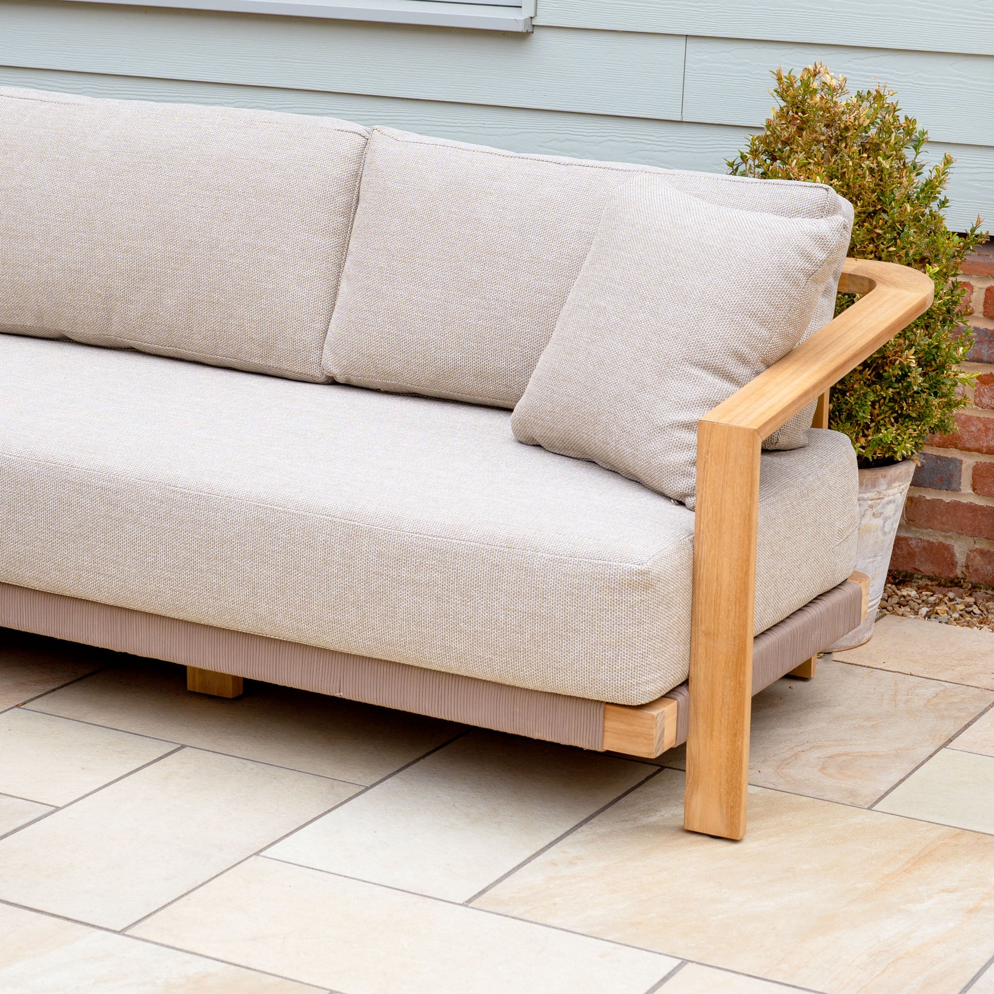 The Tivoli Large Corner Set in Fawn with a marble coffee table features on a tiled patio, beside a potted shrub and brick wall.