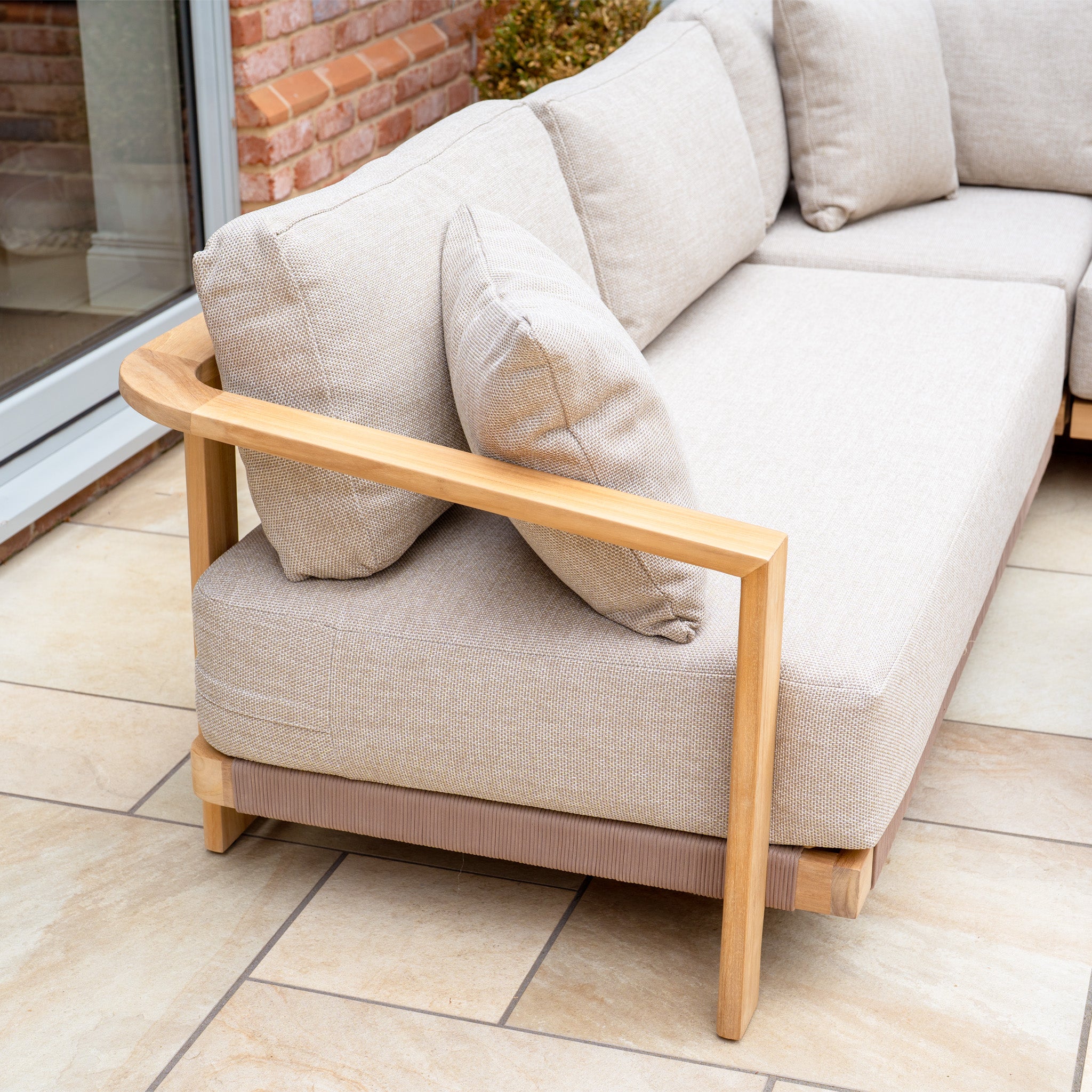 The Tivoli Large Corner Set in Fawn with a marble coffee table features a light teak wood frame and beige cushions, styled on a tiled patio next to a brick wall and glass door.
