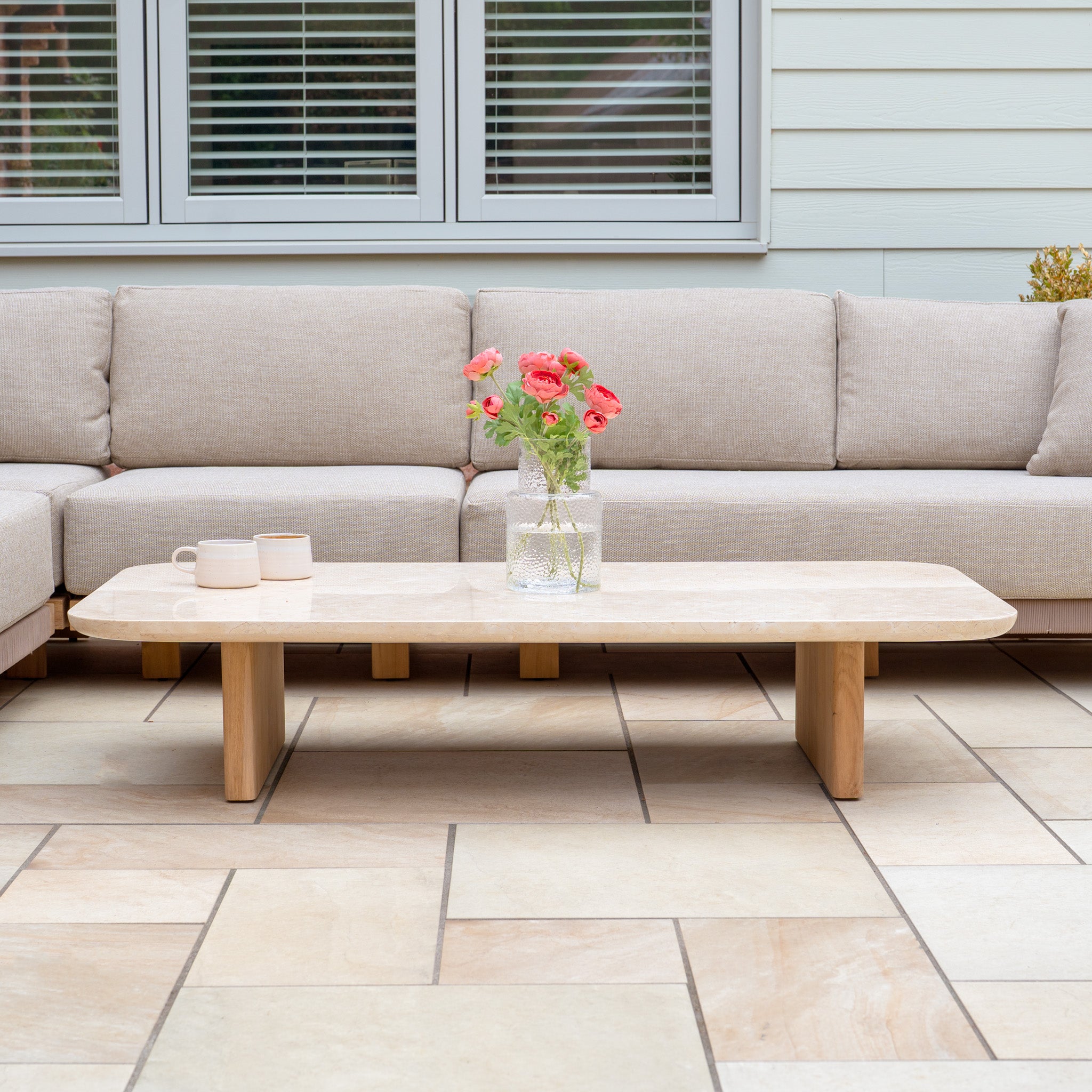 A Tivoli Large Corner Set with Marble Coffee Table in Fawn, featuring pink flowers in a vase and two mugs on a tiled floor.