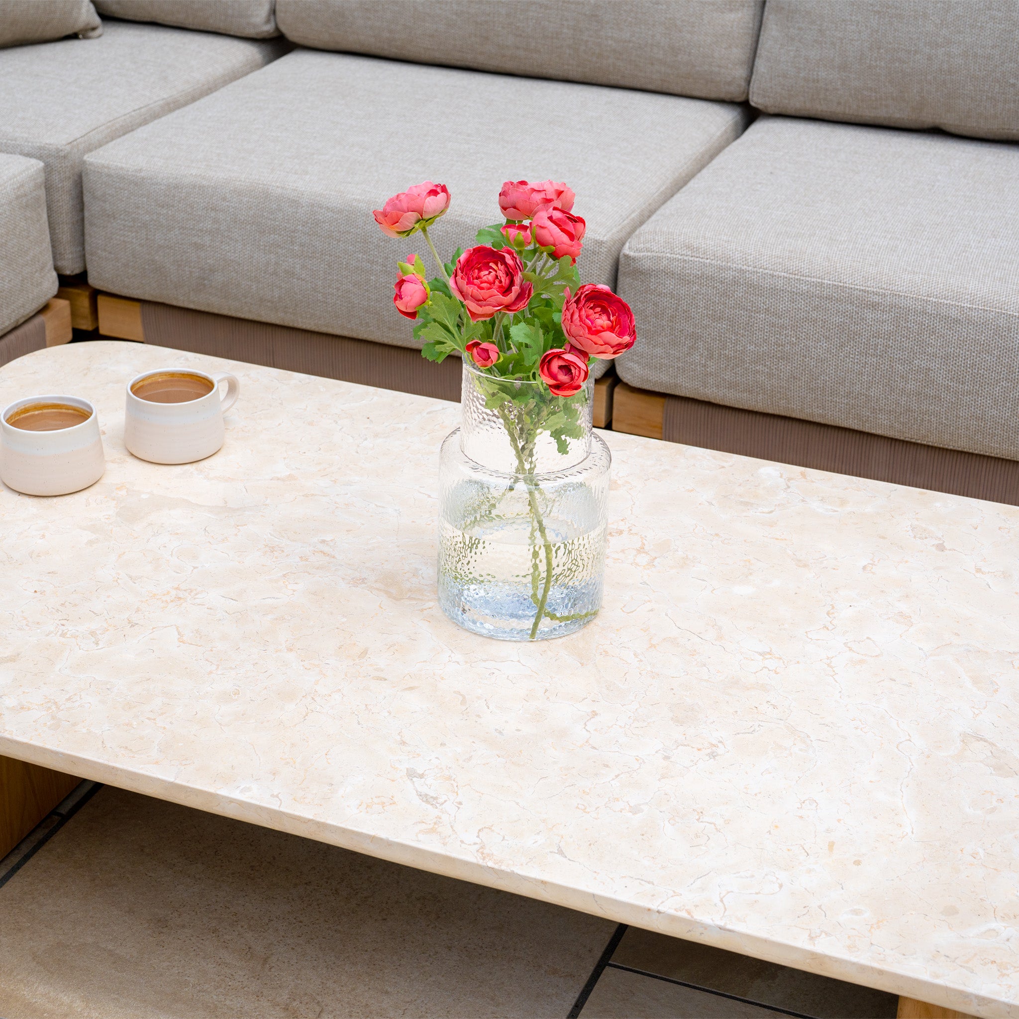 A vase with red flowers and two cups of coffee rest on the Tivoli Large Corner Set with Marble Coffee Table in Fawn, placed before a gray sofa for a cozy atmosphere.