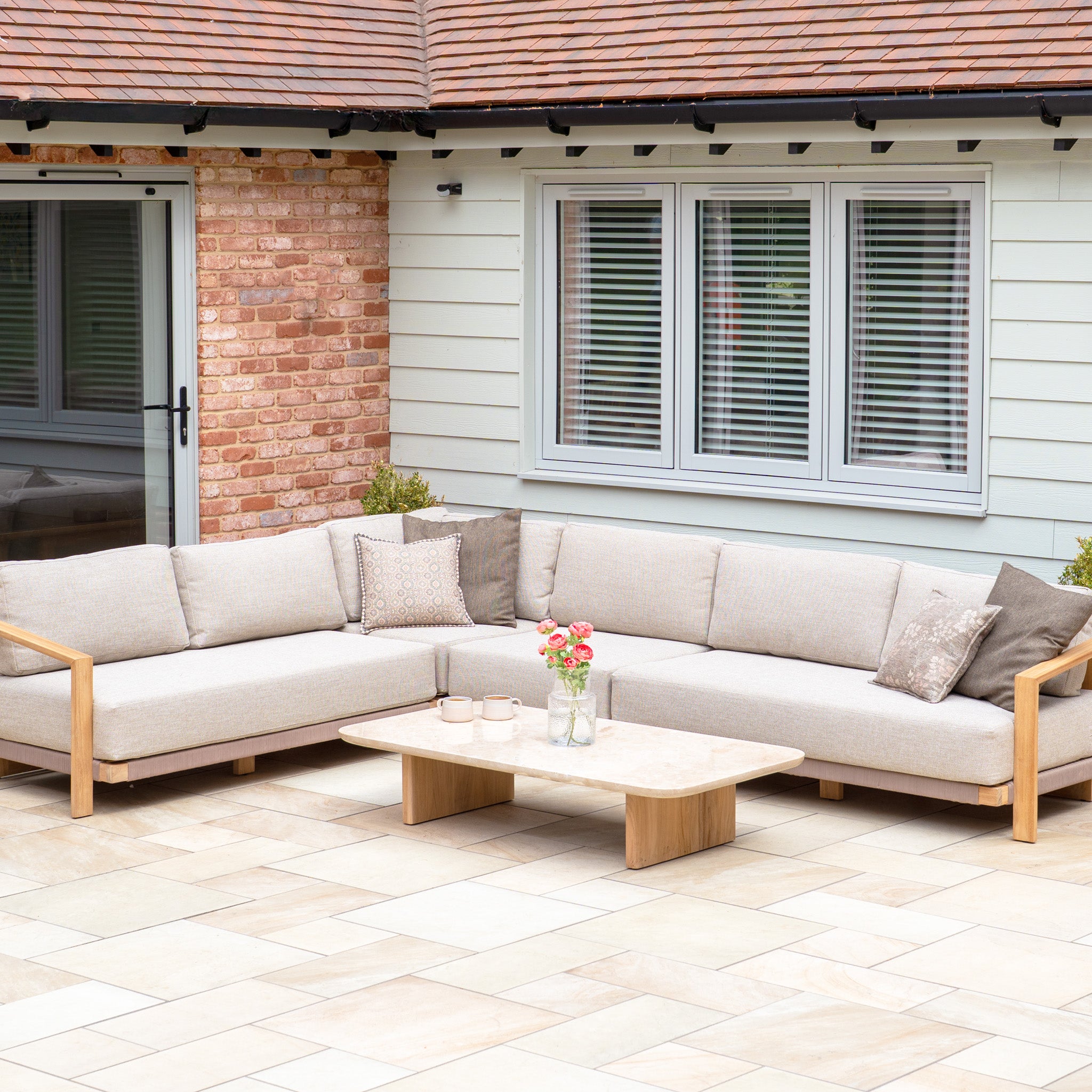 Fawn outdoor sectional sofa from the Tivoli Large Corner Set with cushions and a marble coffee table on a tiled patio beside a house with large windows.