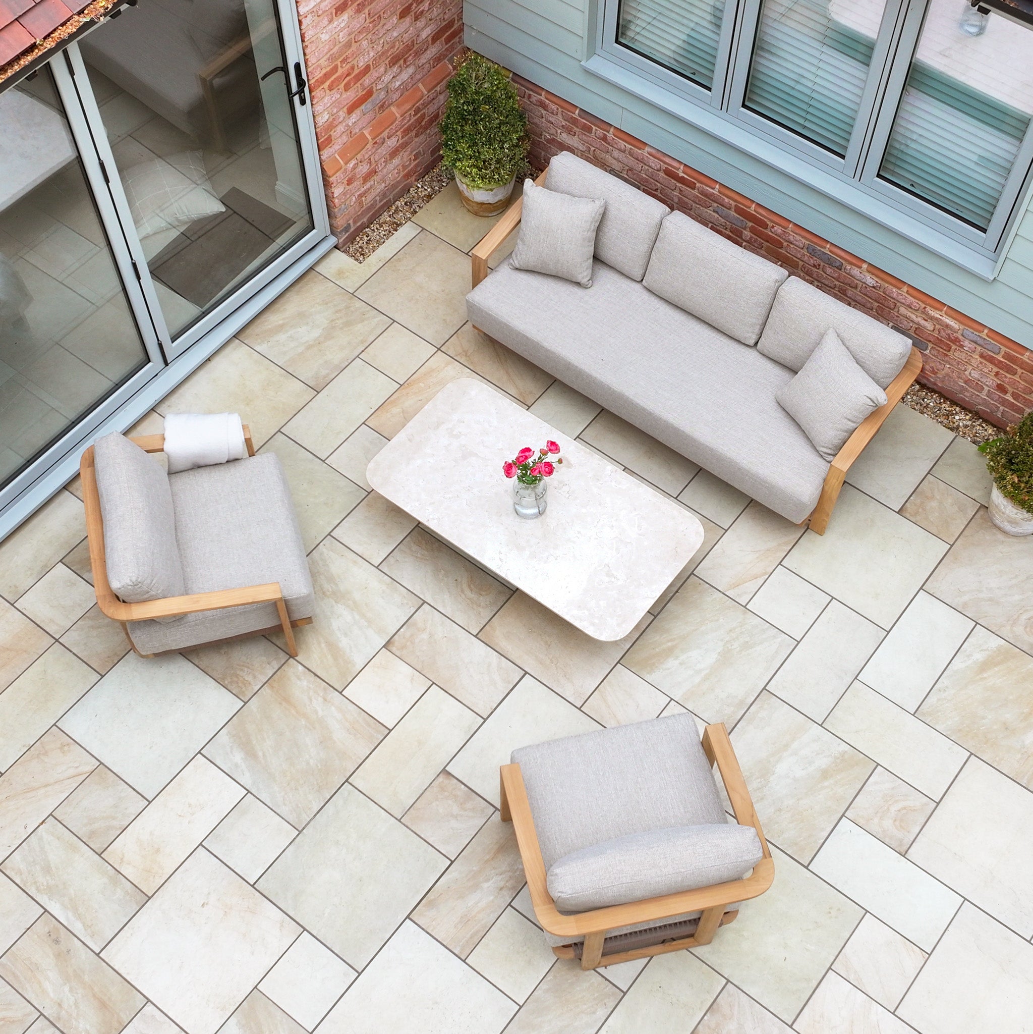 Outdoor patio with the Tivoli 3 Seat Sofa Set with Marble Coffee Table in Fawn, two armchairs, flowers on the table, and potted plants on stone tiles.