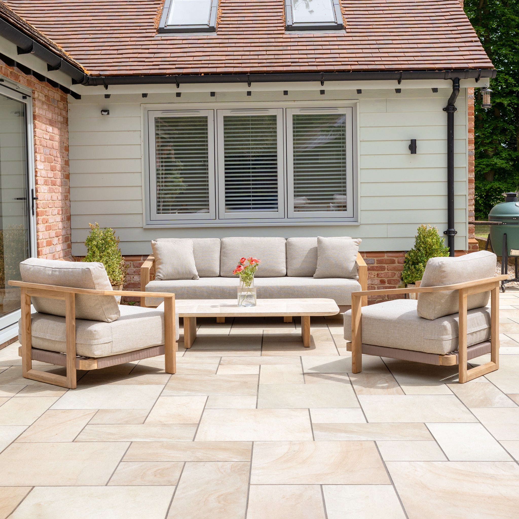 Outdoor patio with a Tivoli 3 Seat Sofa Set with Marble Coffee Table in Fawn, featuring beige cushions, wood design, and a vase of flowers in front of the house.
