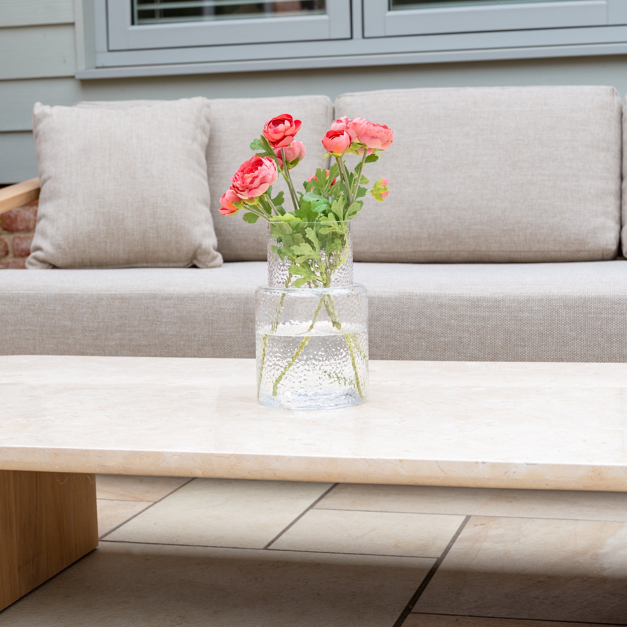 A glass vase with pink flowers sits on the marble coffee table of the Tivoli 3 Seat Sofa Set with Marble Coffee Table in Fawn, creating an inviting outdoor space.