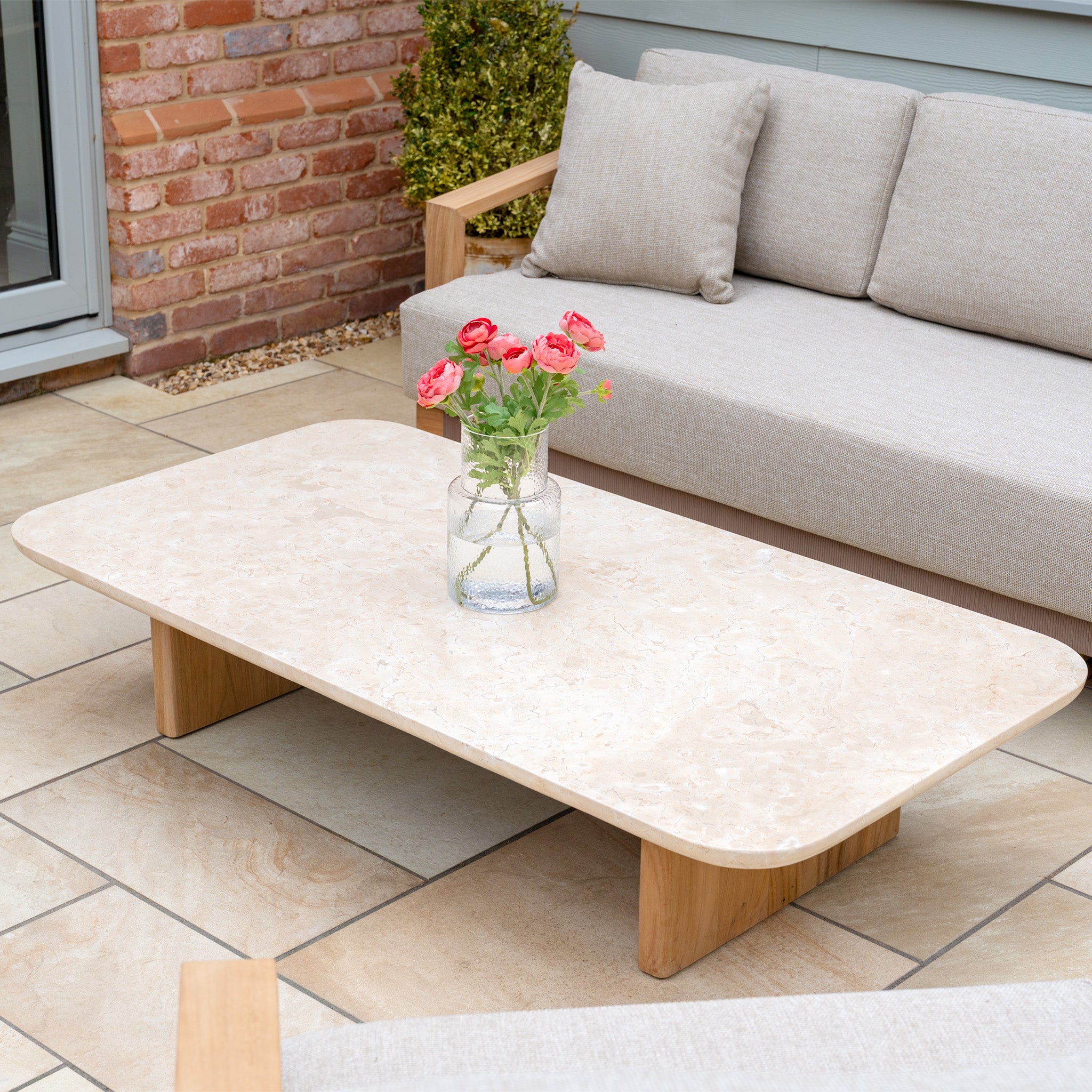 The Tivoli 3 Seat Sofa Set in Fawn features a marble coffee table, complemented by a glass vase with pink flowers, creating an inviting outdoor patio seating area.