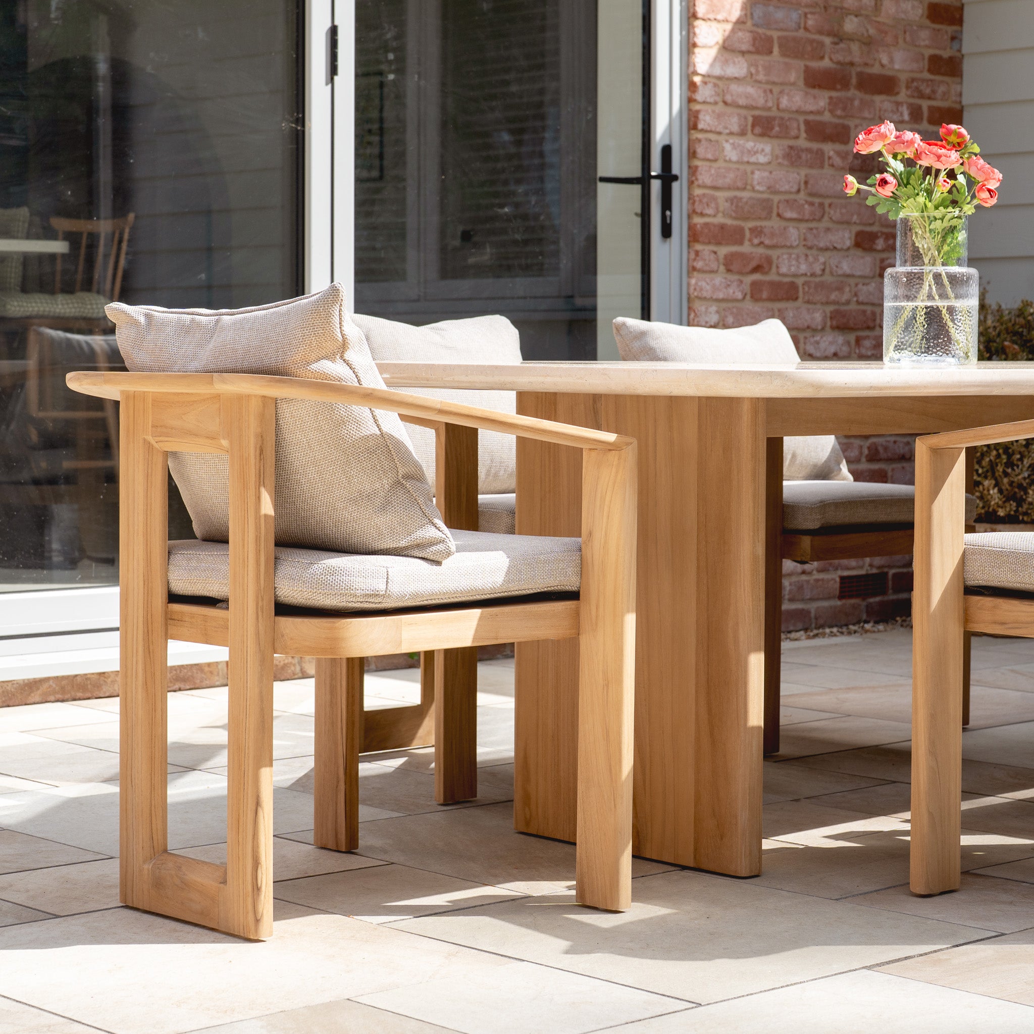 The Tivoli 6 Seat Dining Set features a marble dining table in fawn, elegant teak frame details, beige-cushioned chairs, and understated outdoor luxury by the glass door.