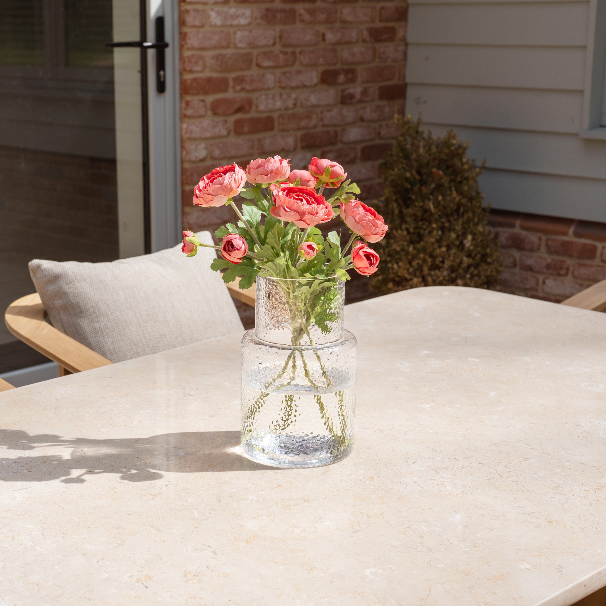 A glass vase with pink flowers sits on the marble dining table of the Tivoli 6 Seat Dining Set in Fawn outdoors, surrounded by teak wood chairs and set beside a brick wall.