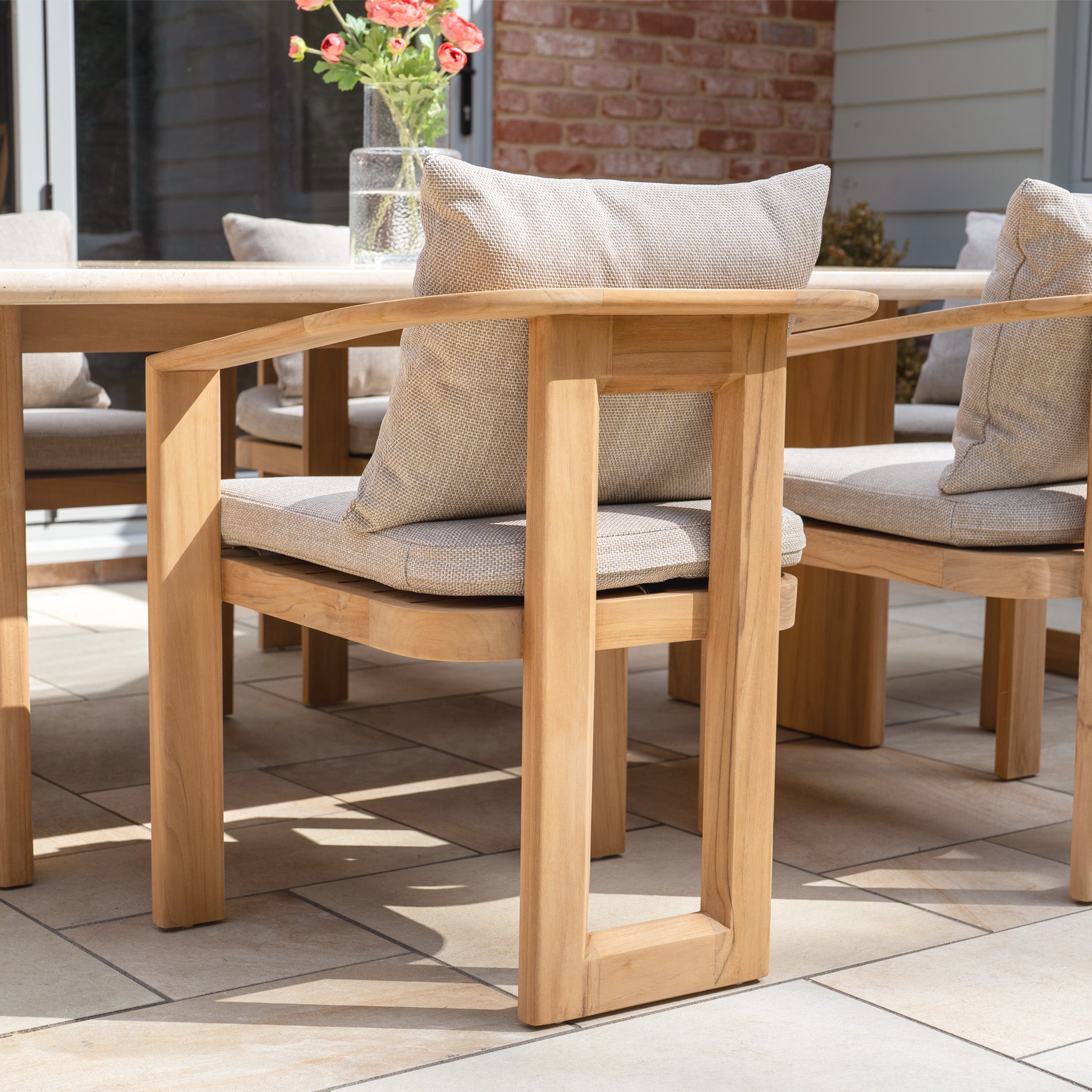 The Tivoli 6 Seat Dining Set with Marble Dining Table in Fawn features teak wood chairs with beige cushions arranged on a tiled patio, creating an inviting outdoor setting.