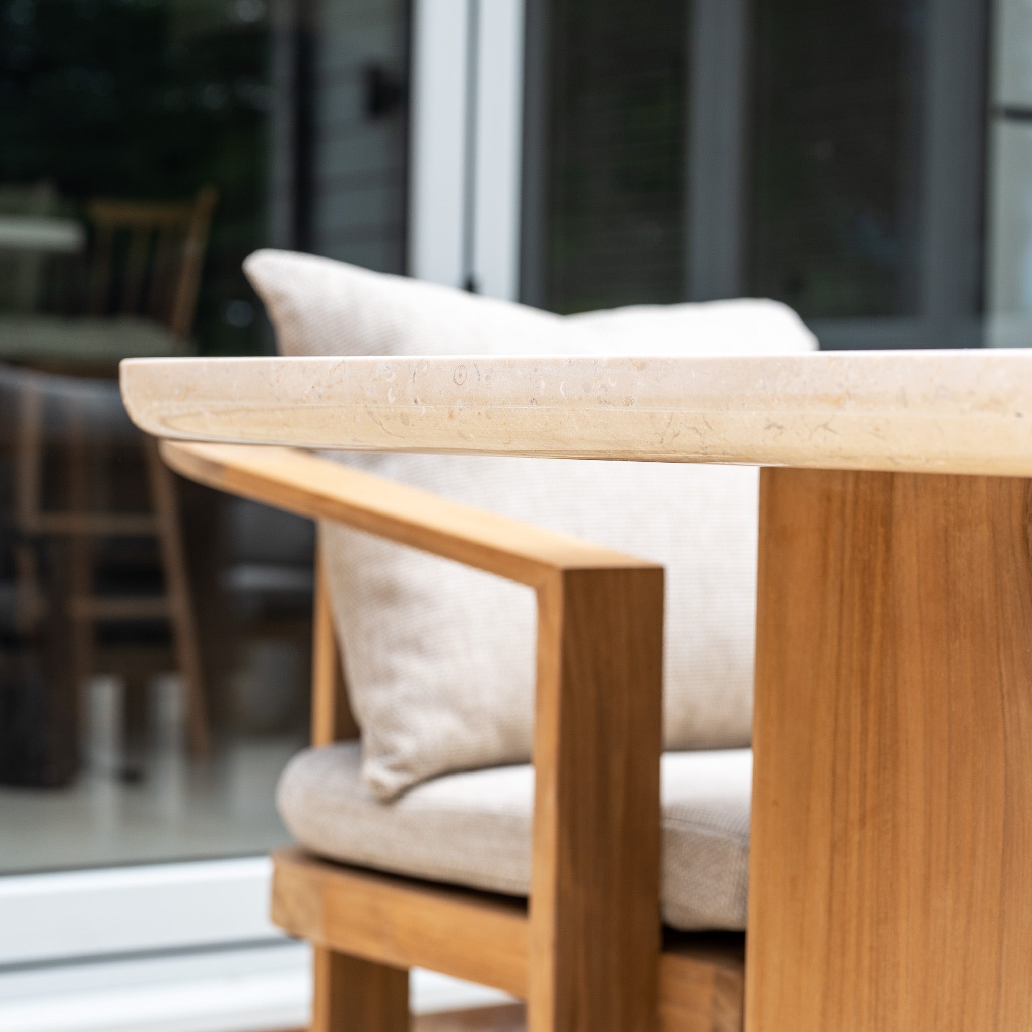Close-up of the Tivoli 6 Seat Dining Set with Marble Dining Table in Fawn, featuring a teak wood chair with a beige cushion, placed outdoors near glass doors.