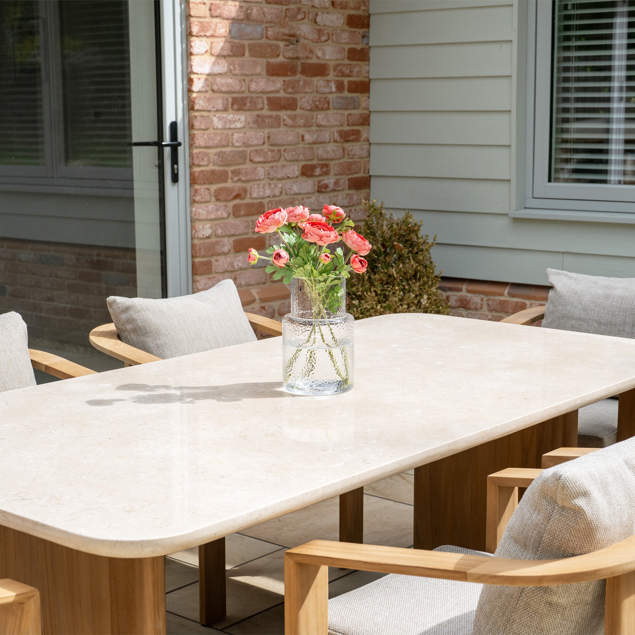 A vase of pink flowers sits on the Tivoli 6 Seat Dining Set with Marble Dining Table in Fawn, surrounded by cushioned wooden chairs for a cozy outdoor dining experience.