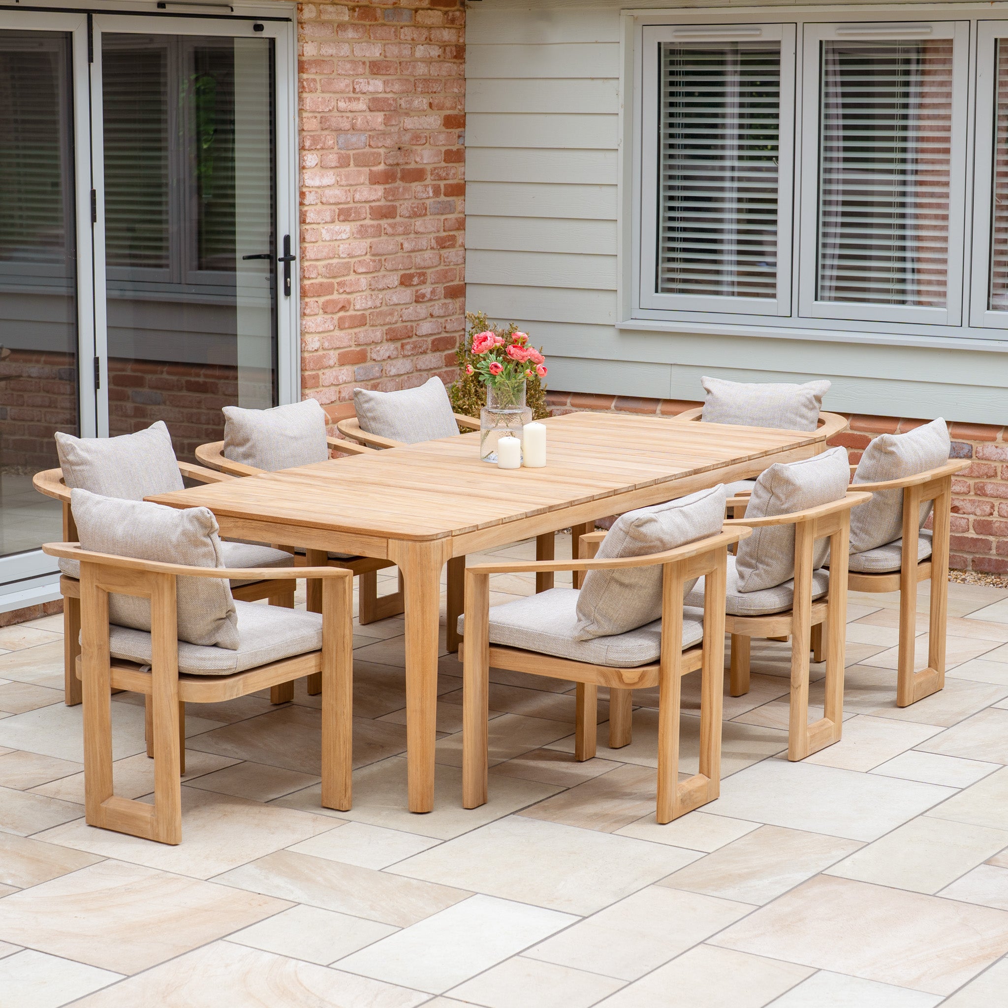 The Tivoli 8 Seat Dining Set with Teak Dining Table in Fawn, shown on a tiled patio near a brick and siding house, features UV-resistant cushions.