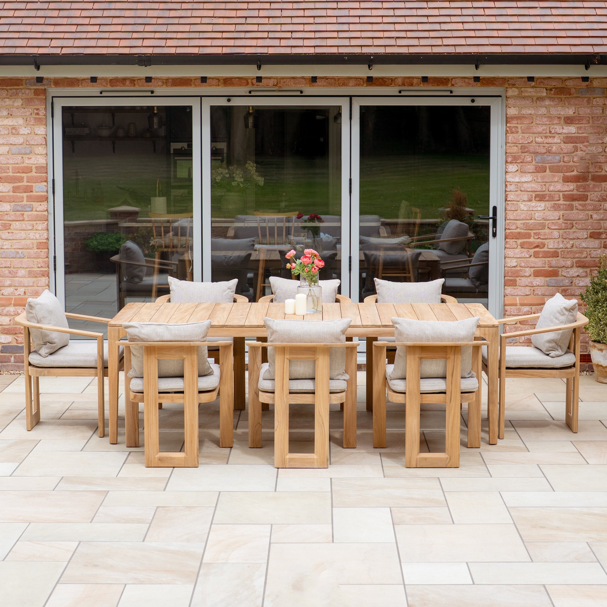 The Tivoli 8 Seat Dining Set with Teak Dining Table in Fawn features UV resistant cushions and is shown arranged on a stone patio before sliding glass doors.