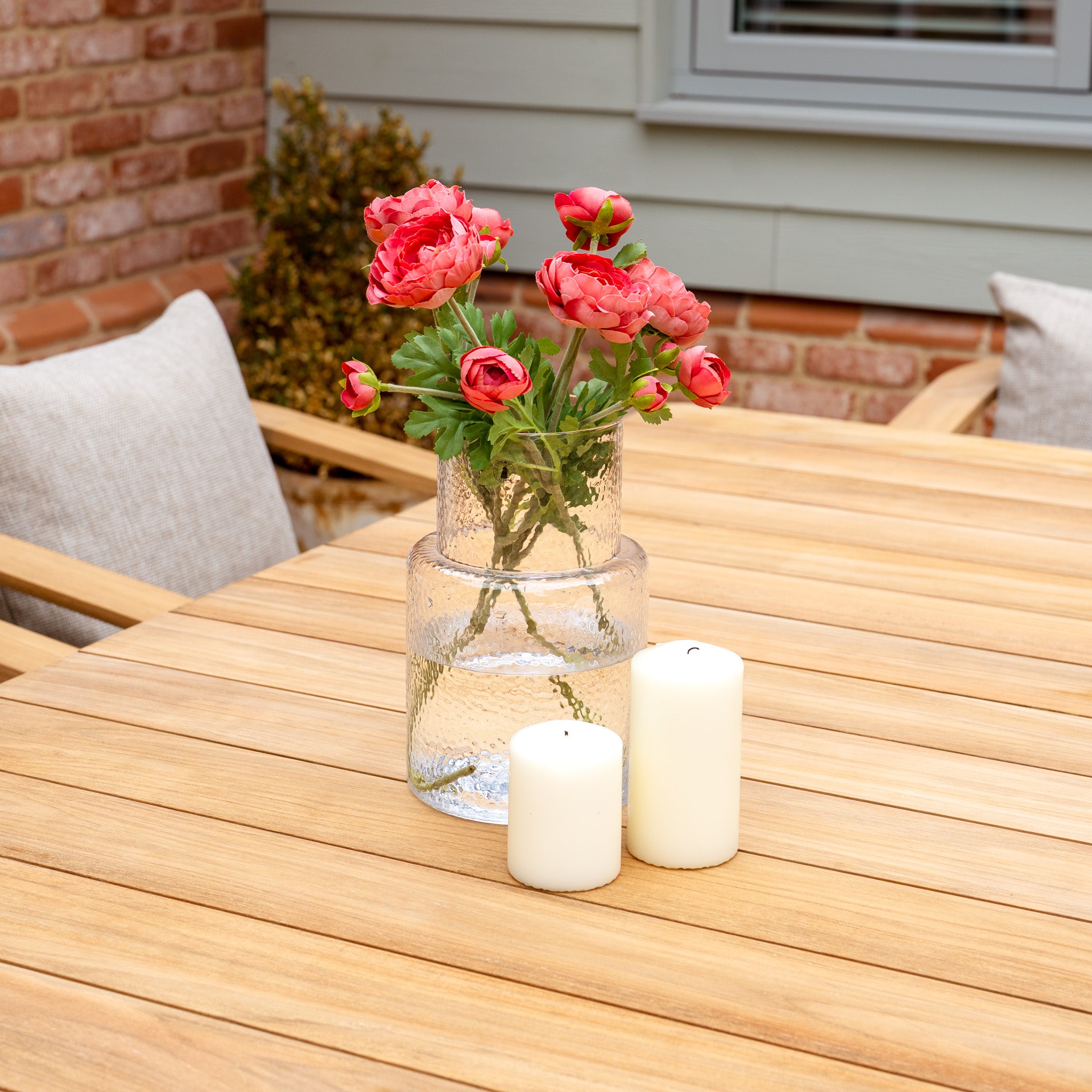 A Tivoli 8 Seat Dining Set with a Teak Dining Table in Fawn is adorned with a clear vase of pink flowers and two white candles, creating a charming outdoor dining atmosphere.