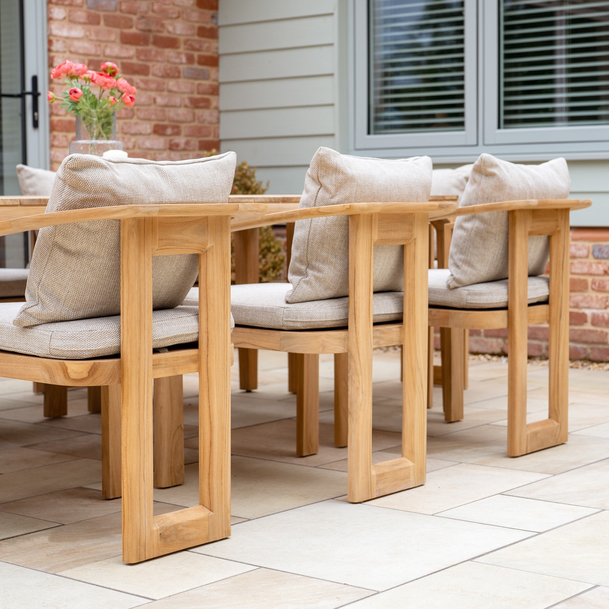The Tivoli 8 Seat Dining Set with Teak Dining Table in Fawn features wooden patio chairs with UV resistant cushions, arranged on a stone-tiled patio near a house for an inviting outdoor dining experience.