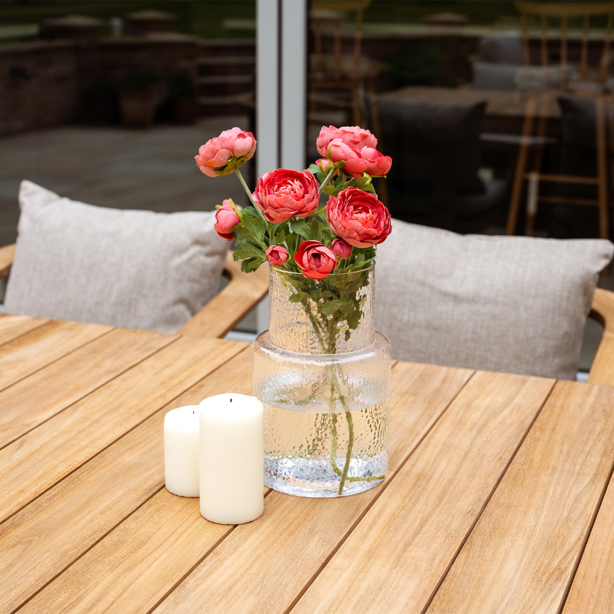 The Tivoli 8 Seat Dining Set with Teak Dining Table in Fawn is styled with a glass vase of pink flowers and two white candles, and features beige UV-resistant cushions for comfortable outdoor dining.