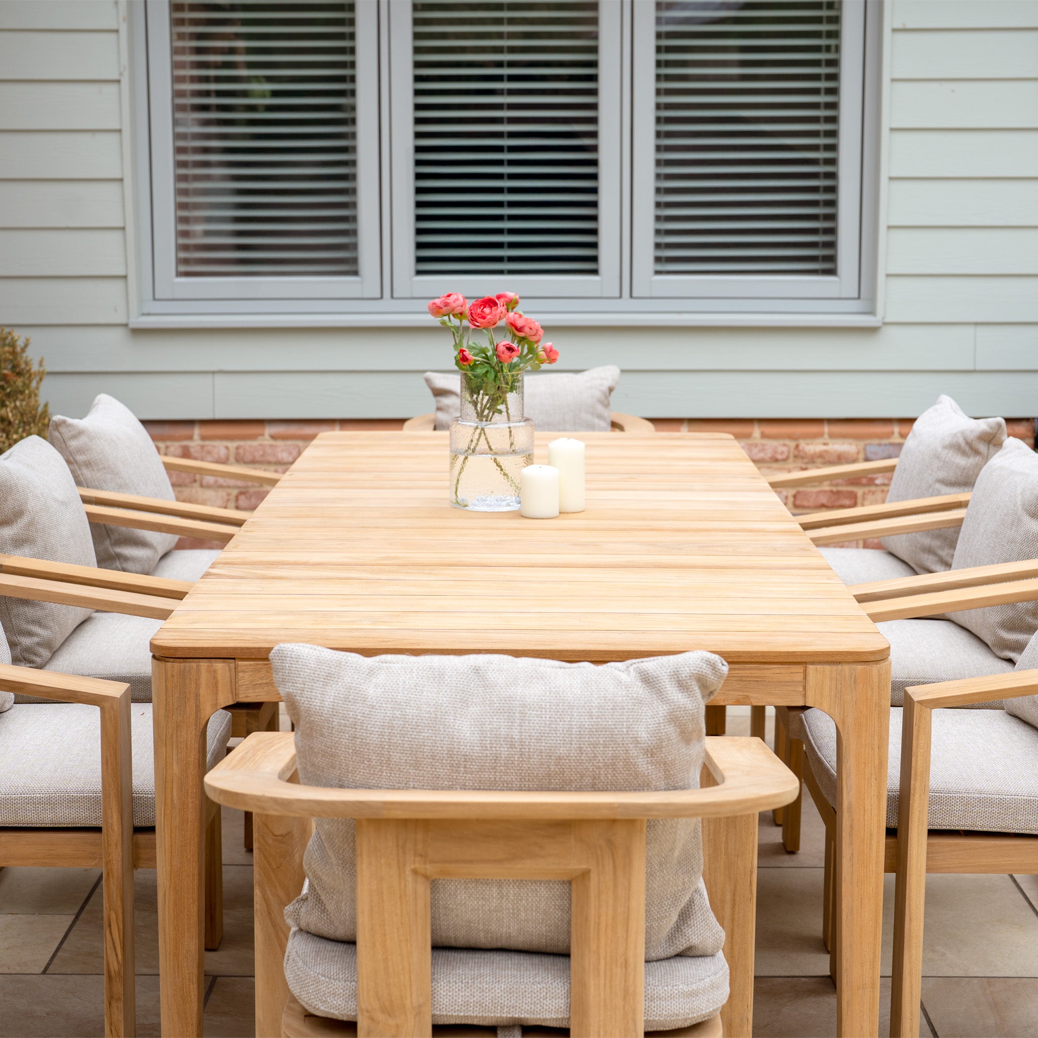 The Tivoli 8 Seat Dining Set with Teak Dining Table in Fawn features UV resistant cushions and is styled with candles and a pink flower vase, creating a stylish centerpiece for outdoor gatherings.