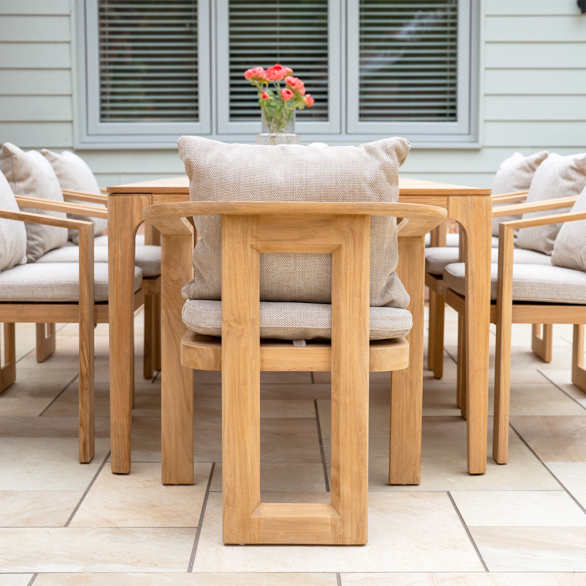 The Tivoli 8 Seat Dining Set with Teak Dining Table in Fawn features patio chairs with UV resistant cushions around a teak table topped with a vase of flowers, set in front of a window.