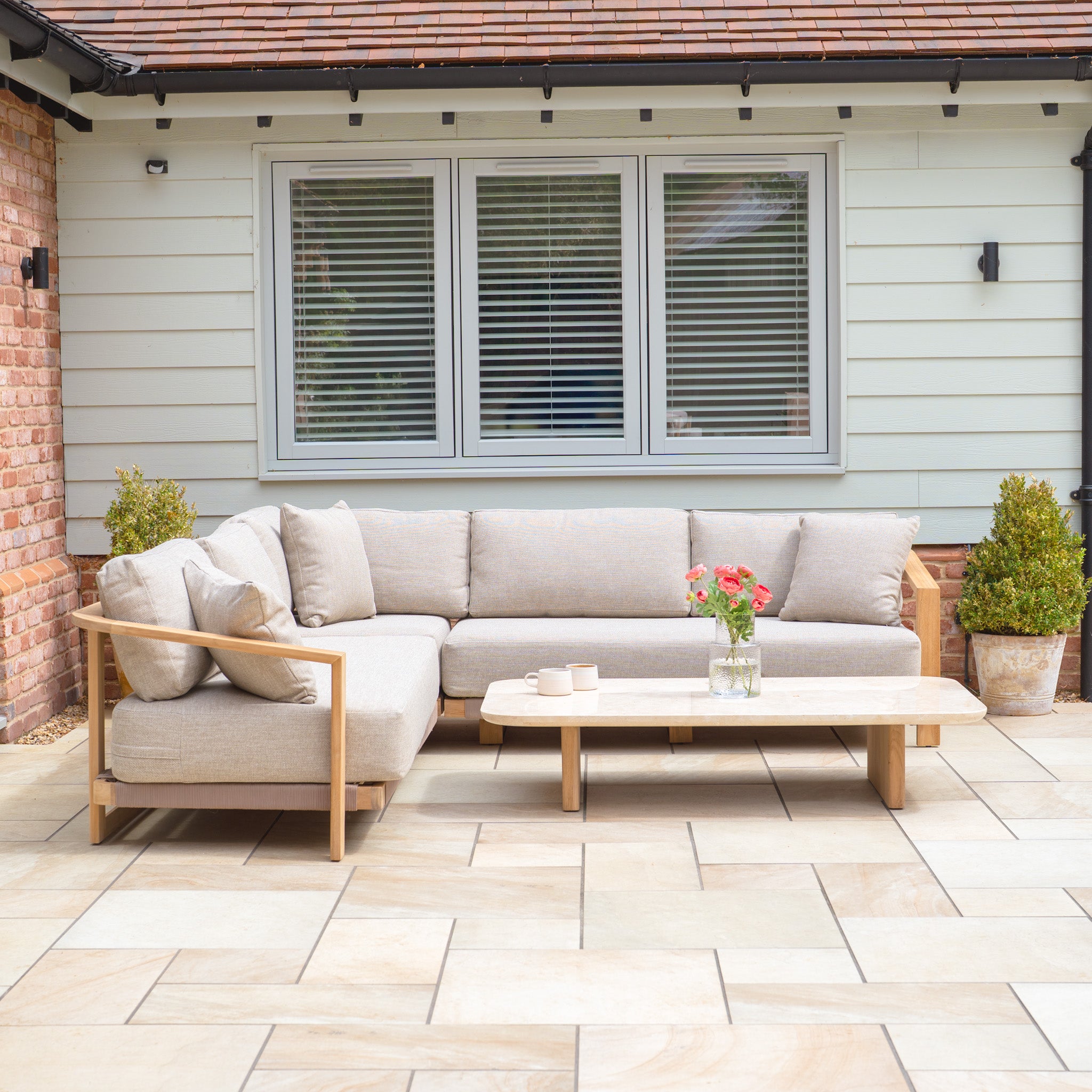 Outdoor patio with a Tivoli Corner Set featuring a fawn L-shaped sofa and marble coffee table, complemented by potted plants in front of a house window.