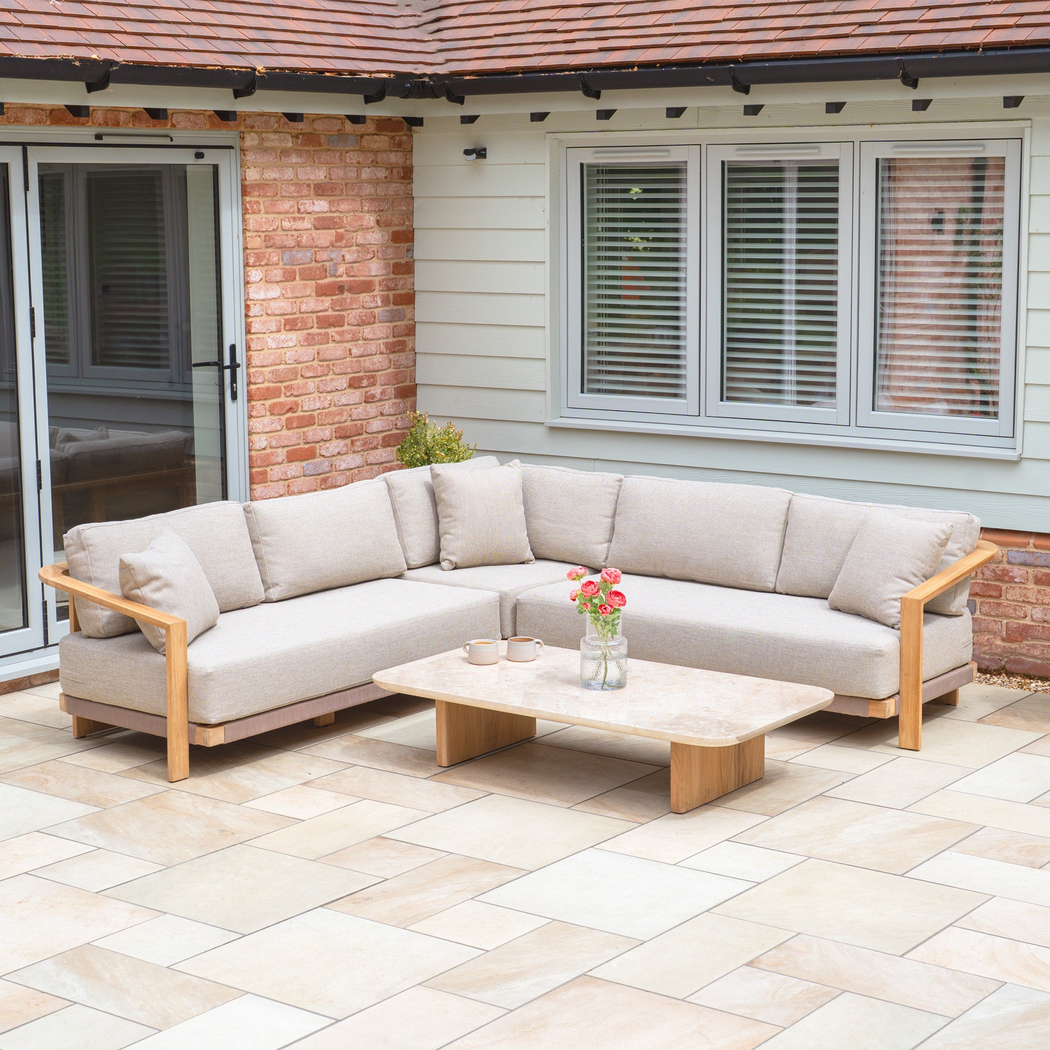 The Tivoli Corner Set with Marble Coffee Table in Fawn features an L-shaped beige outdoor sofa with cushions and a marble coffee table on a tiled patio displaying flowers.
