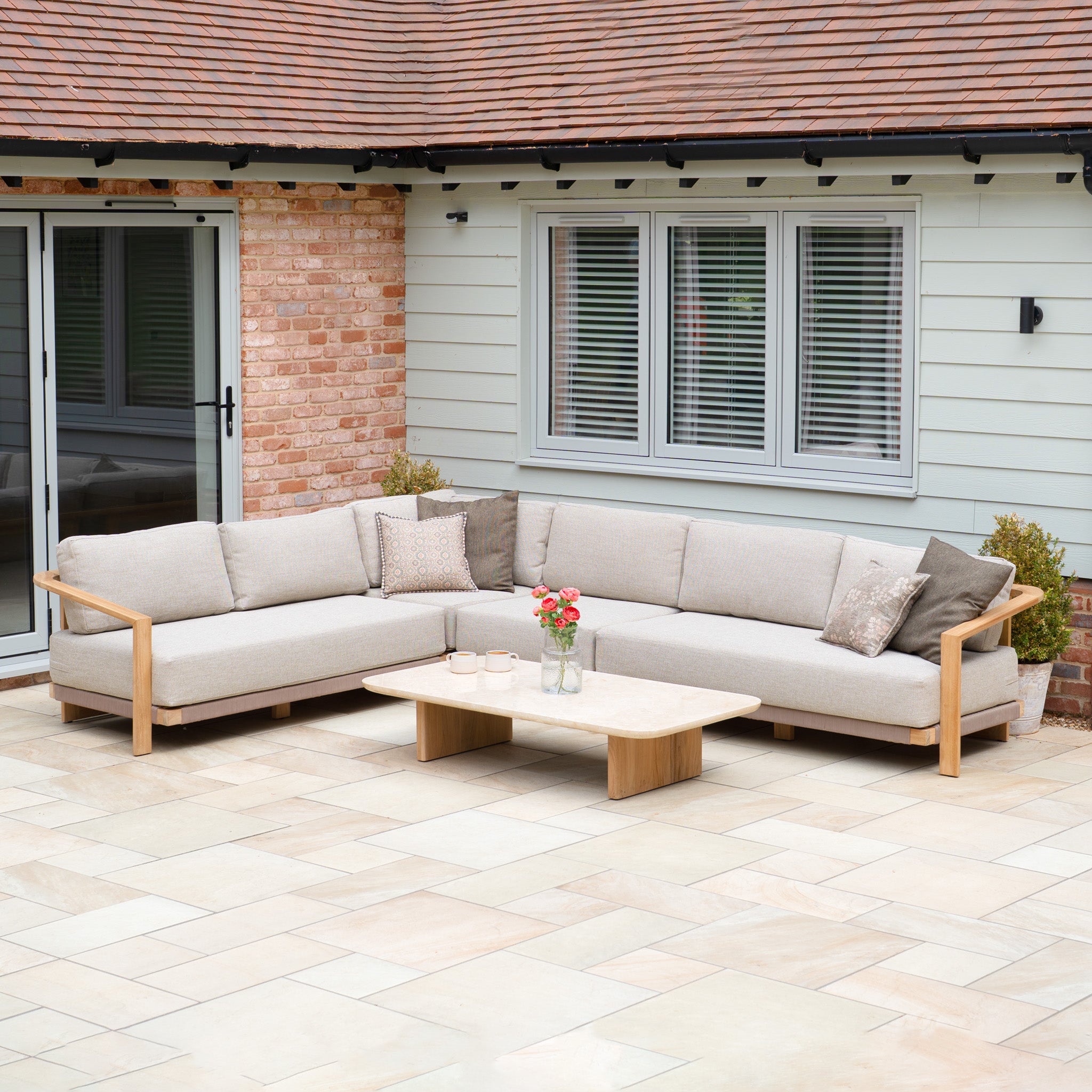 Outdoor patio with the Tivoli Large Corner Set in Fawn, an L-shaped beige sofa and marble coffee table, arranged near flowers outside a house with large windows.
