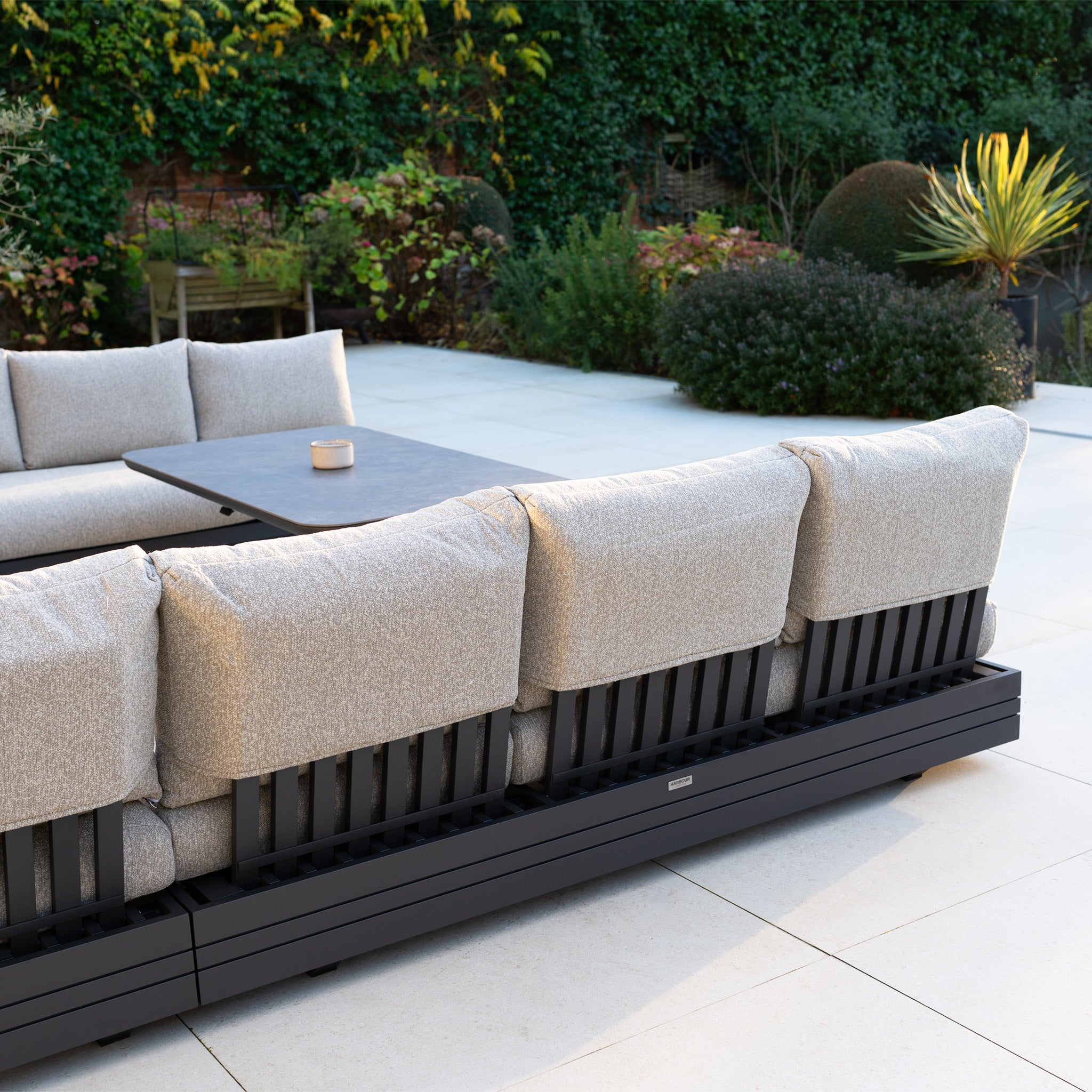 Outdoor patio with the Savona Luxury Outdoor Large U-Shape Corner Sofa Set in Truffle and an Altura Rising Table, surrounded by green plants. The modular garden furniture is crafted from rust-proof aluminium.