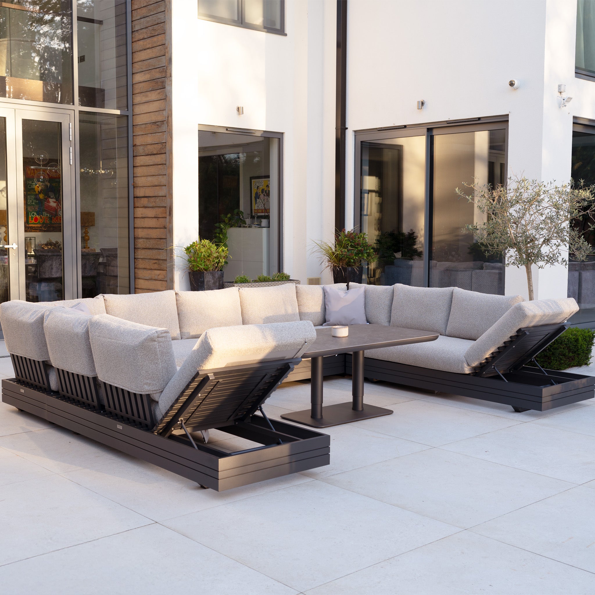 Modern patio featuring the Savona Luxury Outdoor Large U-Shape Corner Sofa Set with Altura Rising Table in Truffle and potted plants. This rust-proof aluminium modular set adds style and durability in front of a white building.