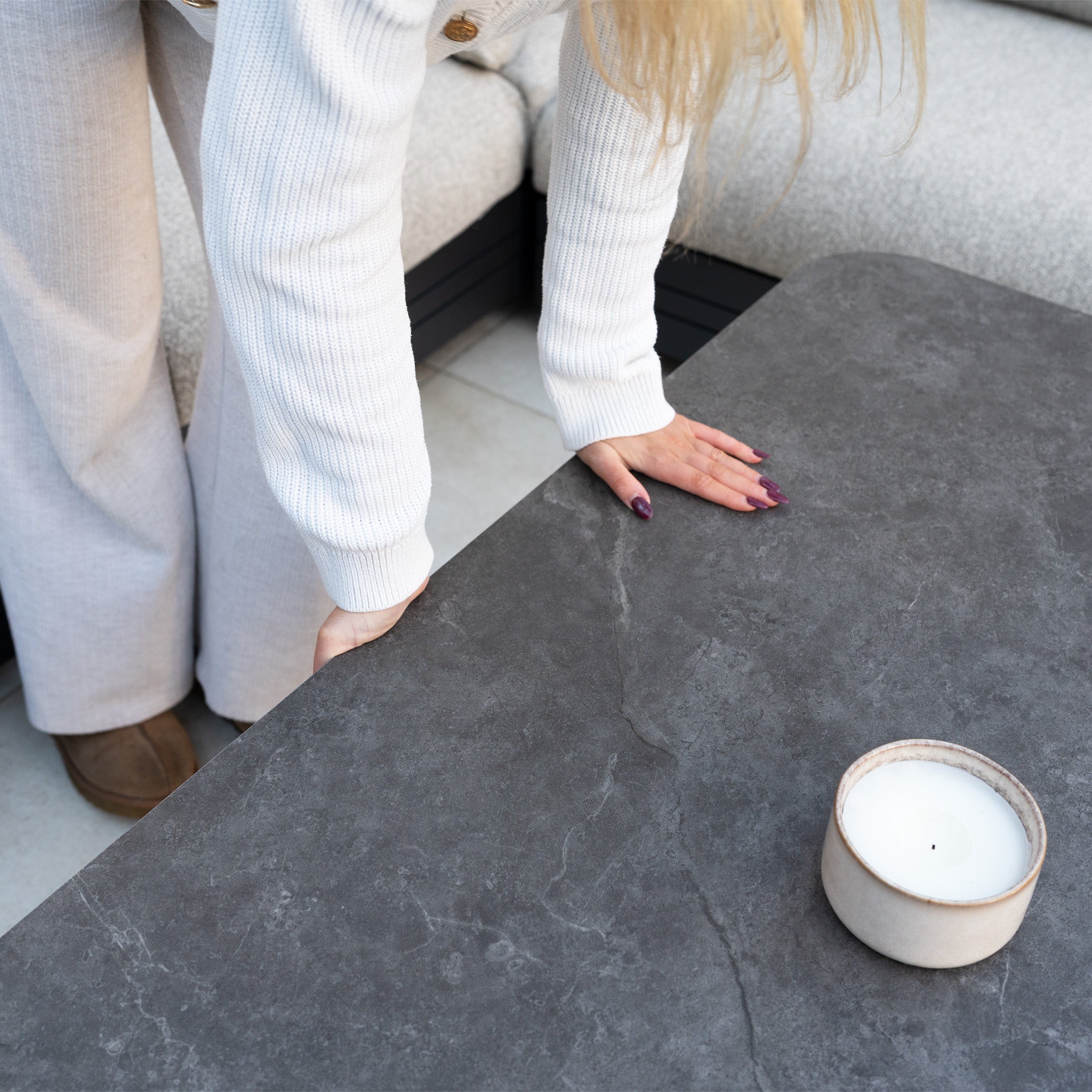 A woman in light clothing leans over a dark stone Altura Rising Table paired with the Savona Luxury Outdoor Large U-Shape Corner Sofa Set in Truffle, featuring rust-proof aluminium modular garden furniture and a white candle in a ceramic holder.