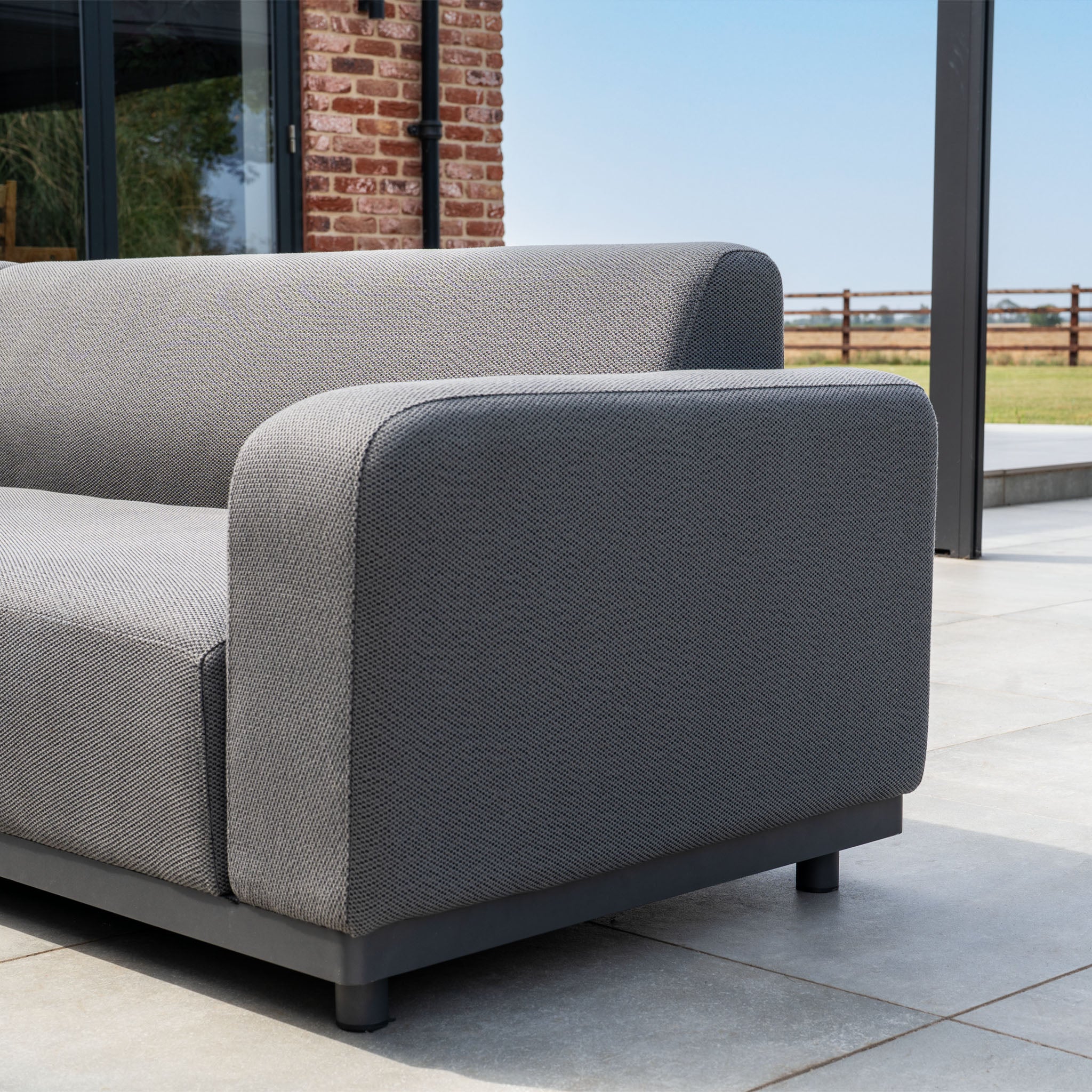 The Luna U-Shape Outdoor Fabric Sofa Set with Altura Rising Table in Grey features a modern aluminium frame, perfect for patios with brick and wood backdrops.