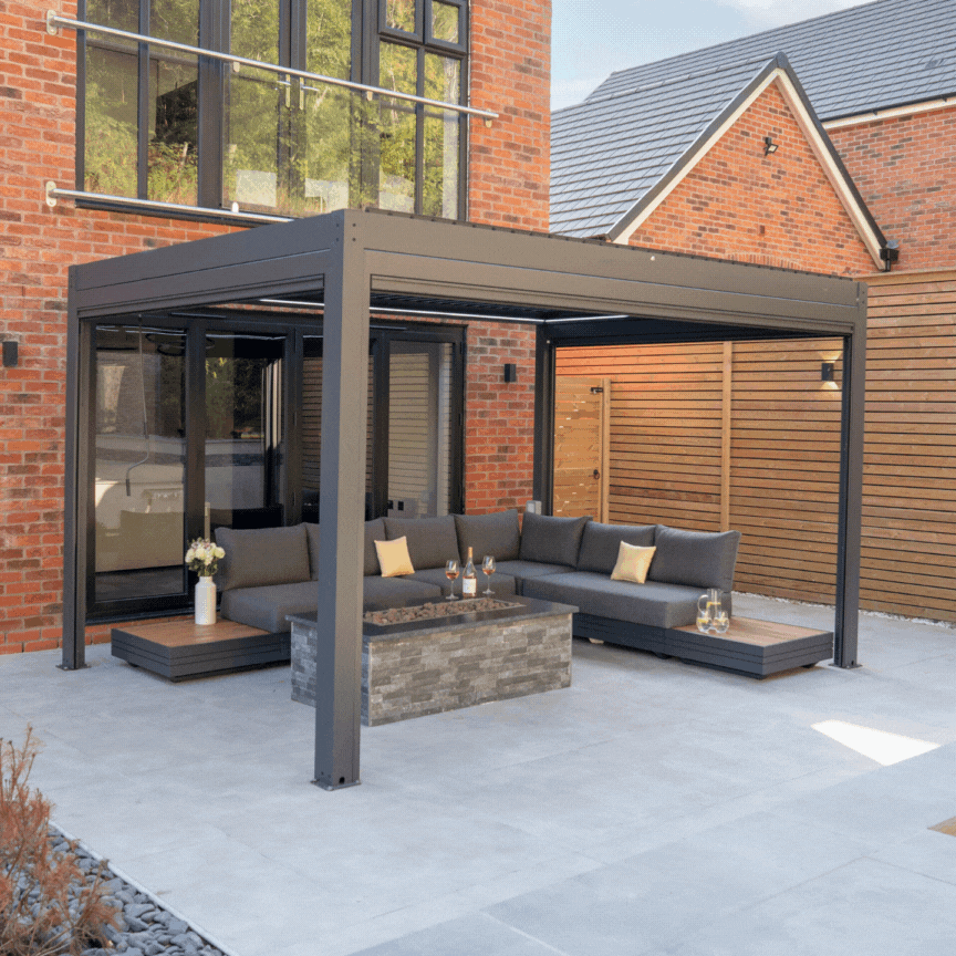 A stylish patio boasts the PergoSTET Plus 3m x 4m Grey Pergola, featuring three drop sides and LED lighting, complemented by a sectional sofa and coffee table against the brick house backdrop for the perfect outdoor seating arrangement.