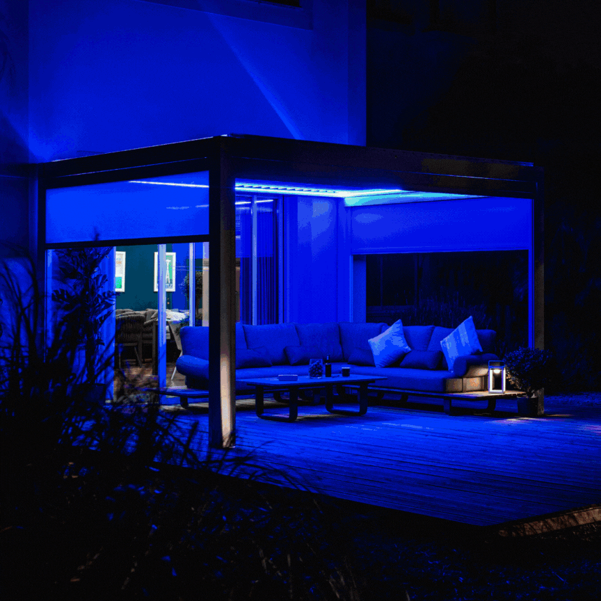 A modern patio with blue lighting showcases a sectional sofa and wooden deck at night, elegantly enhanced by the PergoSTET Plus 3m x 4m Rectangular Pergola in grey, featuring three drop sides and LED lighting for added sophistication and style to your outdoor oasis.