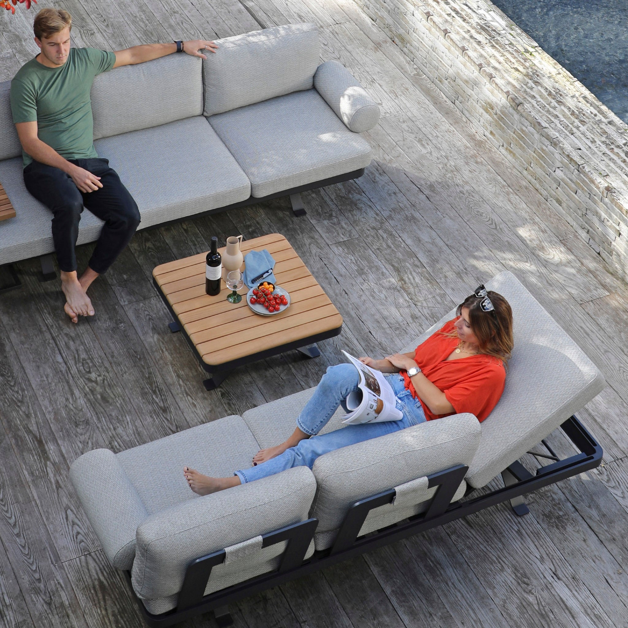 Two people relax on a comprehensive and elegant Vienna Corner Group Set in Grey on a wooden deck; one reads a magazine, while the other sits nearby, enjoying their outdoor living space.