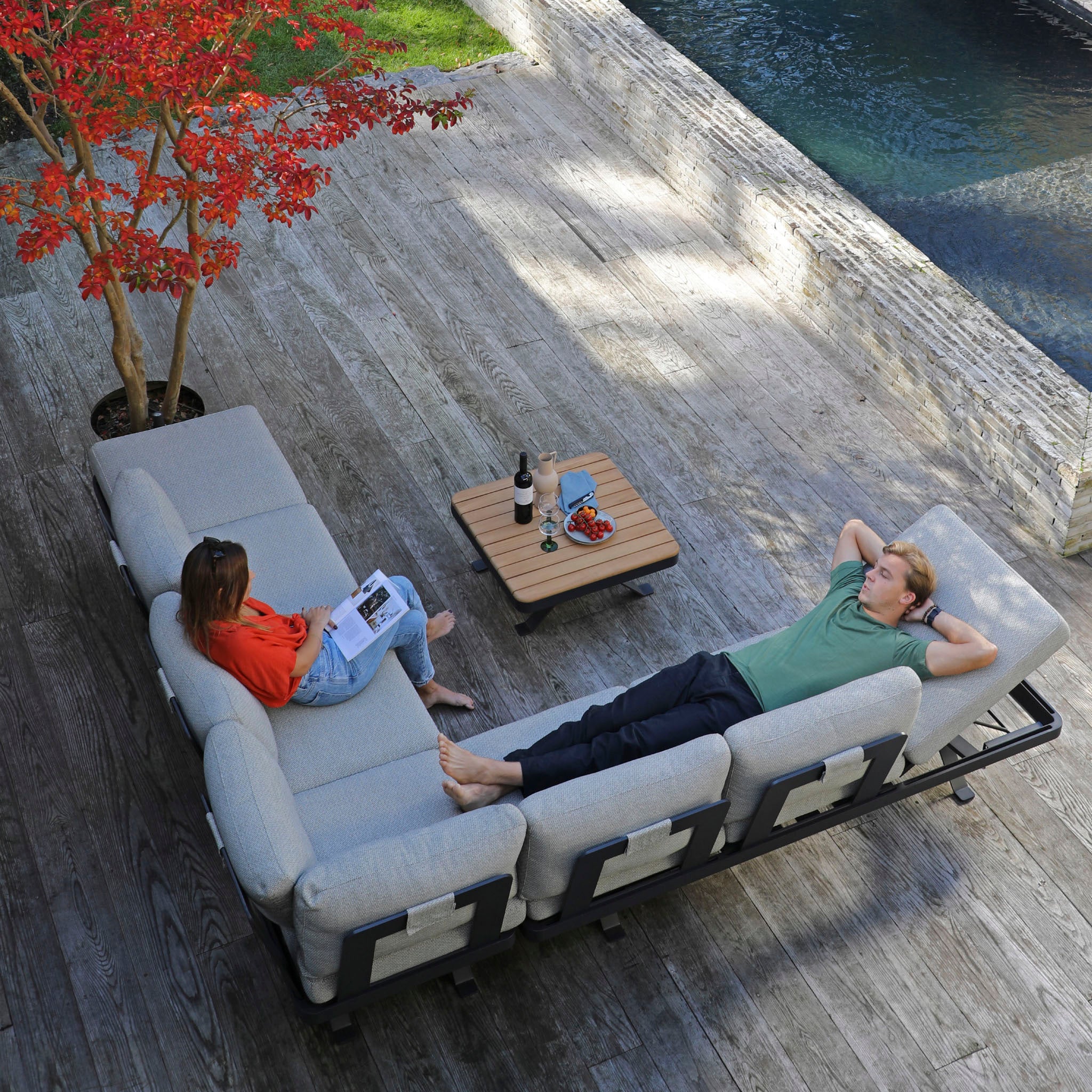 Two people relaxing on an outdoor sofa by a pool, one reading and the other lying down with hands behind the head. The Vienna Corner Group Set in Grey they’re enjoying is crafted from high-end materials, making it perfect for stylish outdoor living.