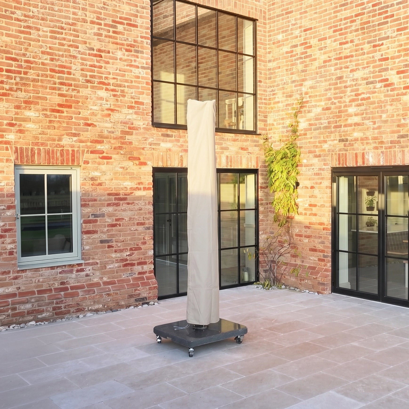A tall patio heater with a beige cover stands on a tiled patio beside the Volos 3m x 3m Square Cantilever Parasol with LED Lighting and a 120kg granite base in slate, in front of a brick building with large windows.