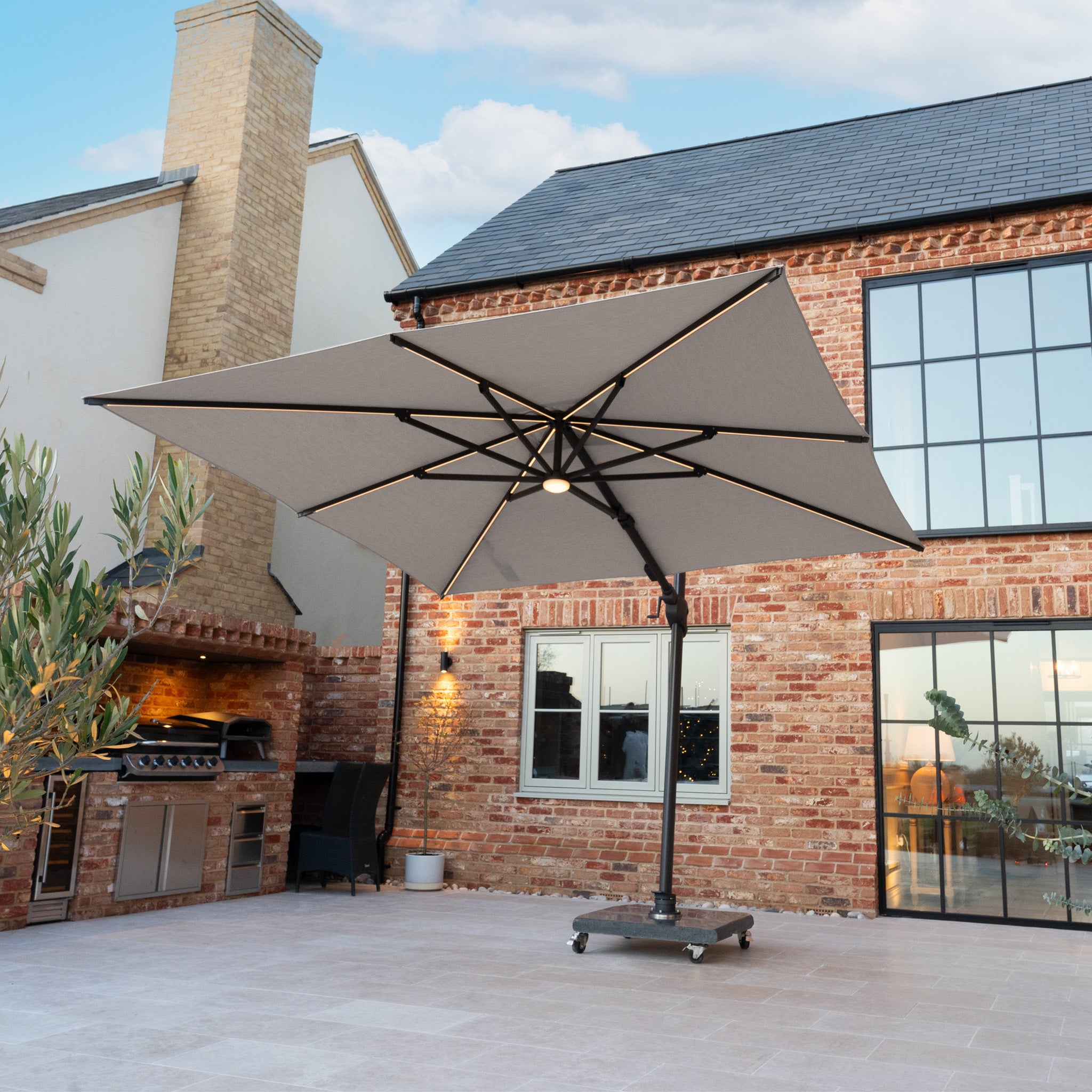 The Volos 3m x 3m Square Cantilever Parasol with LED Lighting and 120kg Granite Base in Slate stands on a spacious tiled patio next to a brick house and outdoor kitchen.