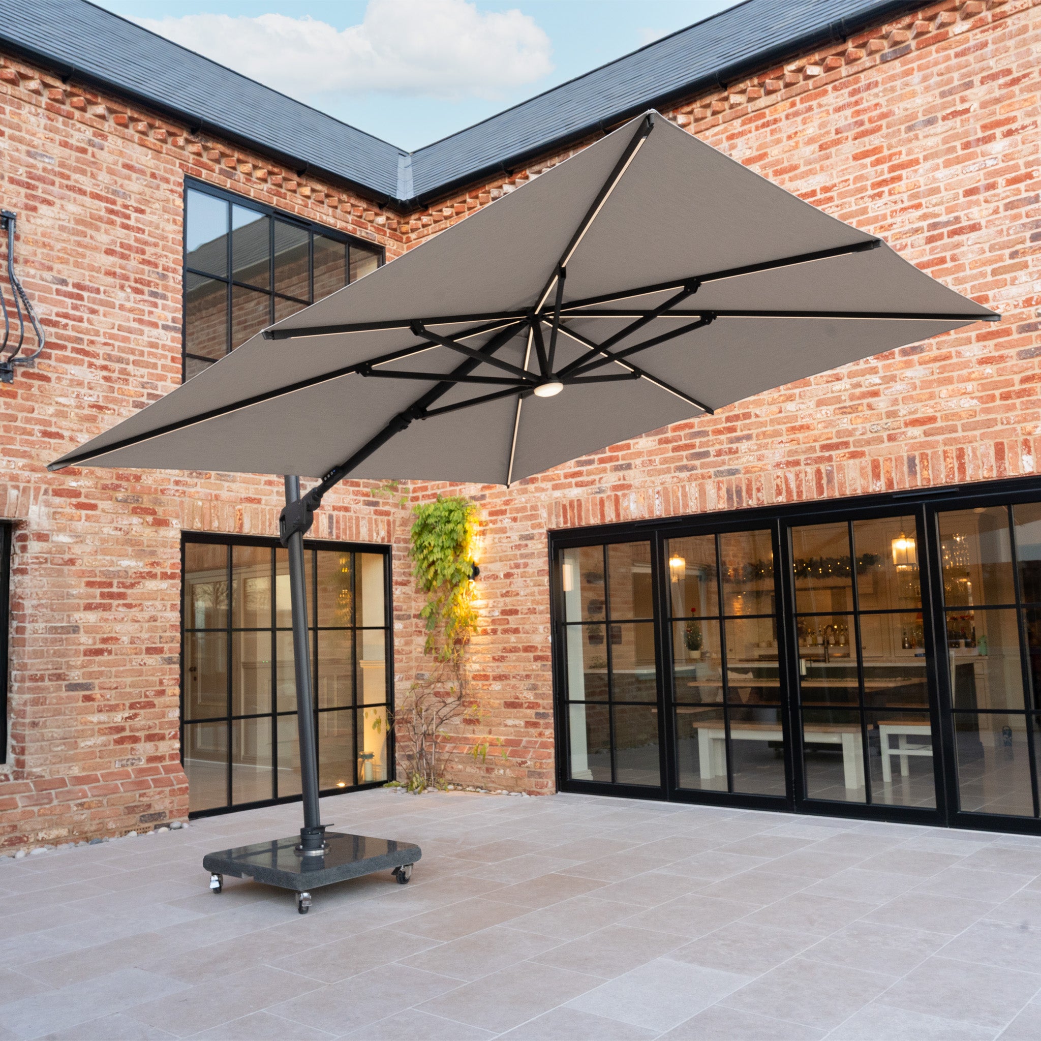 The Volos 3m x 3m Square Cantilever Parasol with LED Lighting and 120kg granite base in Slate stands on wheels in a brick courtyard with glass doors and a tiled floor.