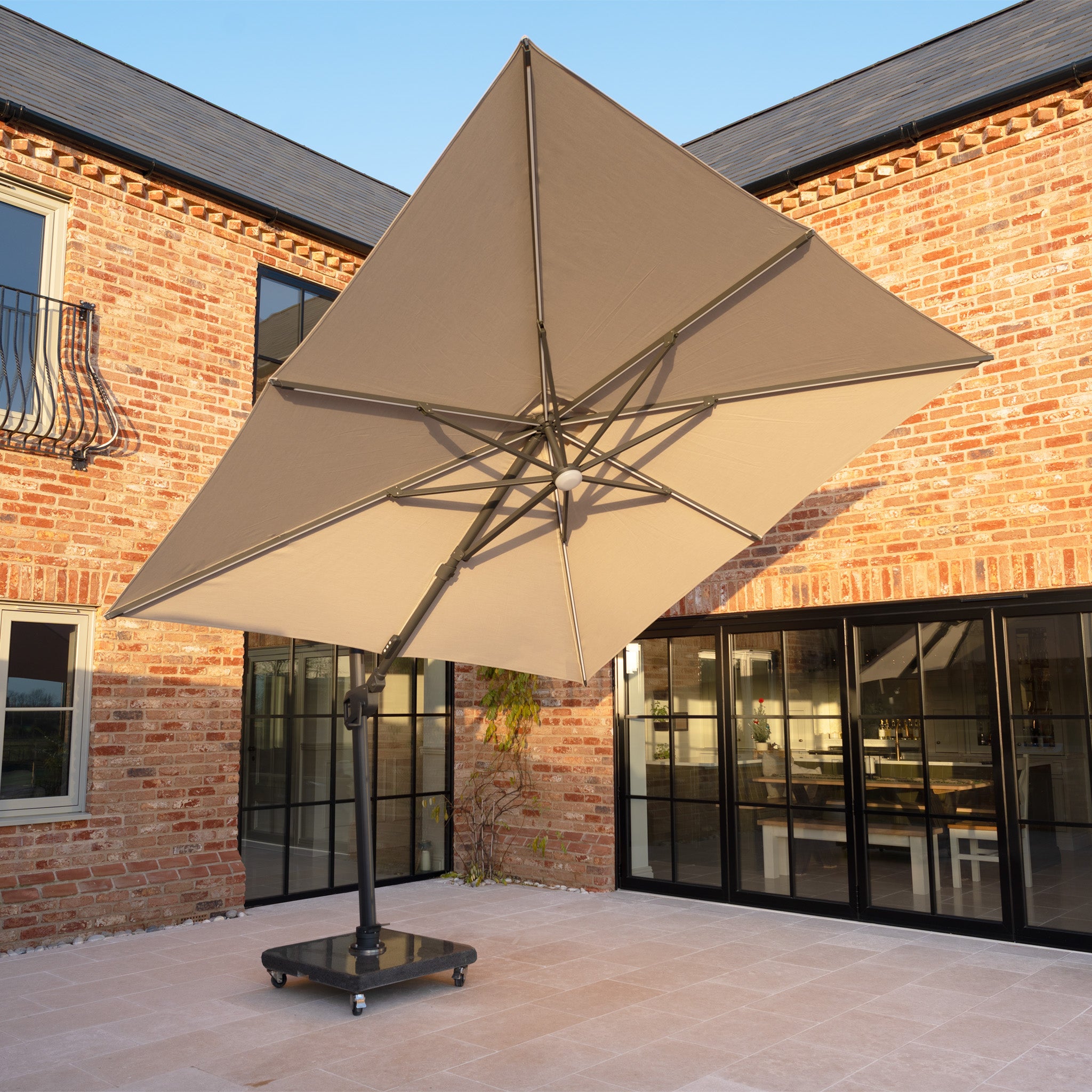 The Volos 3m x 3m Square Cantilever Parasol in Wheat with LED lighting and a 120kg granite base stands on a tiled patio outside a modern brick house with glass doors.