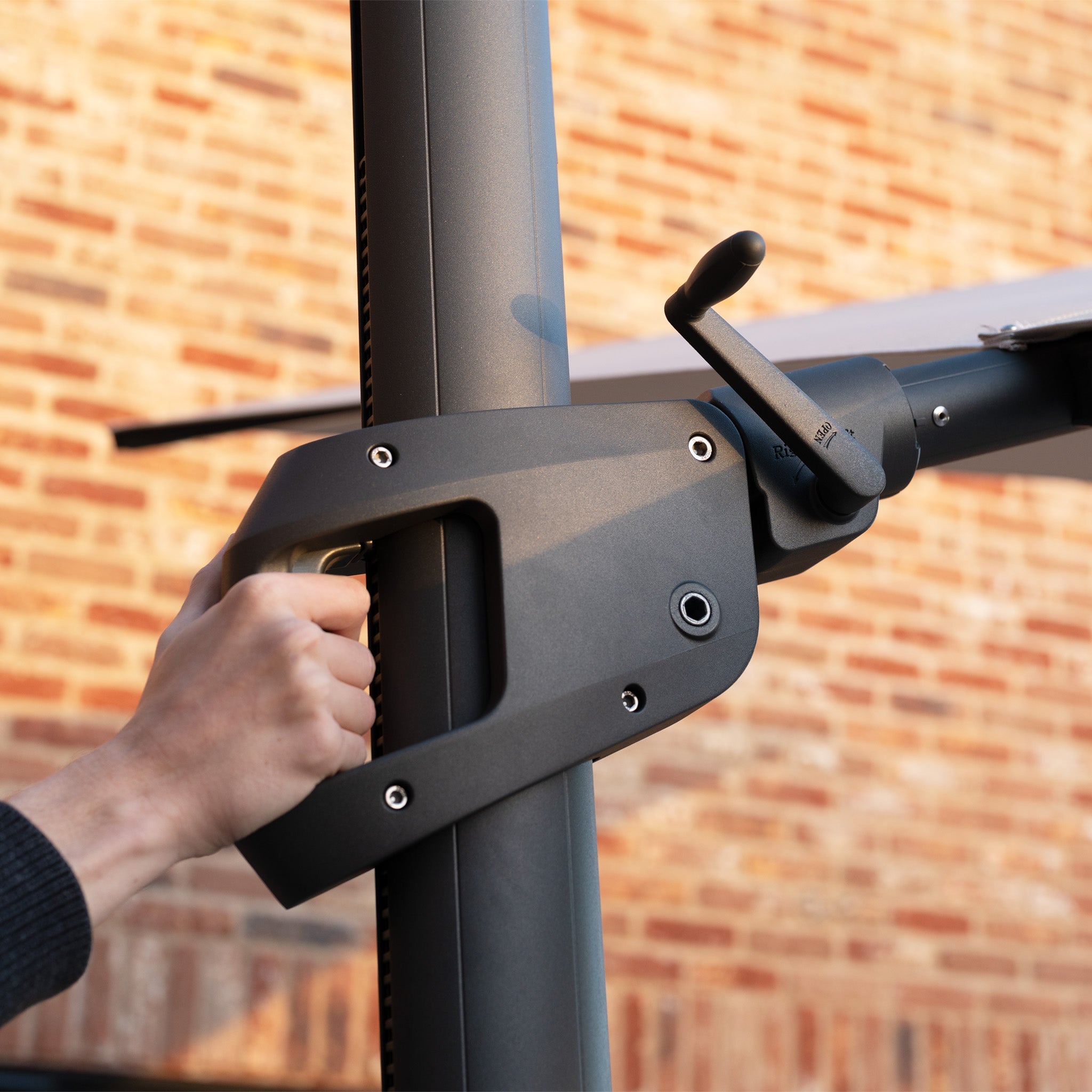 Hand adjusting the crank handle on the Volos 3m x 3m Square Cantilever Parasol in Wheat, featuring a UV-resistant canopy, LED lighting, and a sturdy 120kg granite base, with a brick wall in the background.